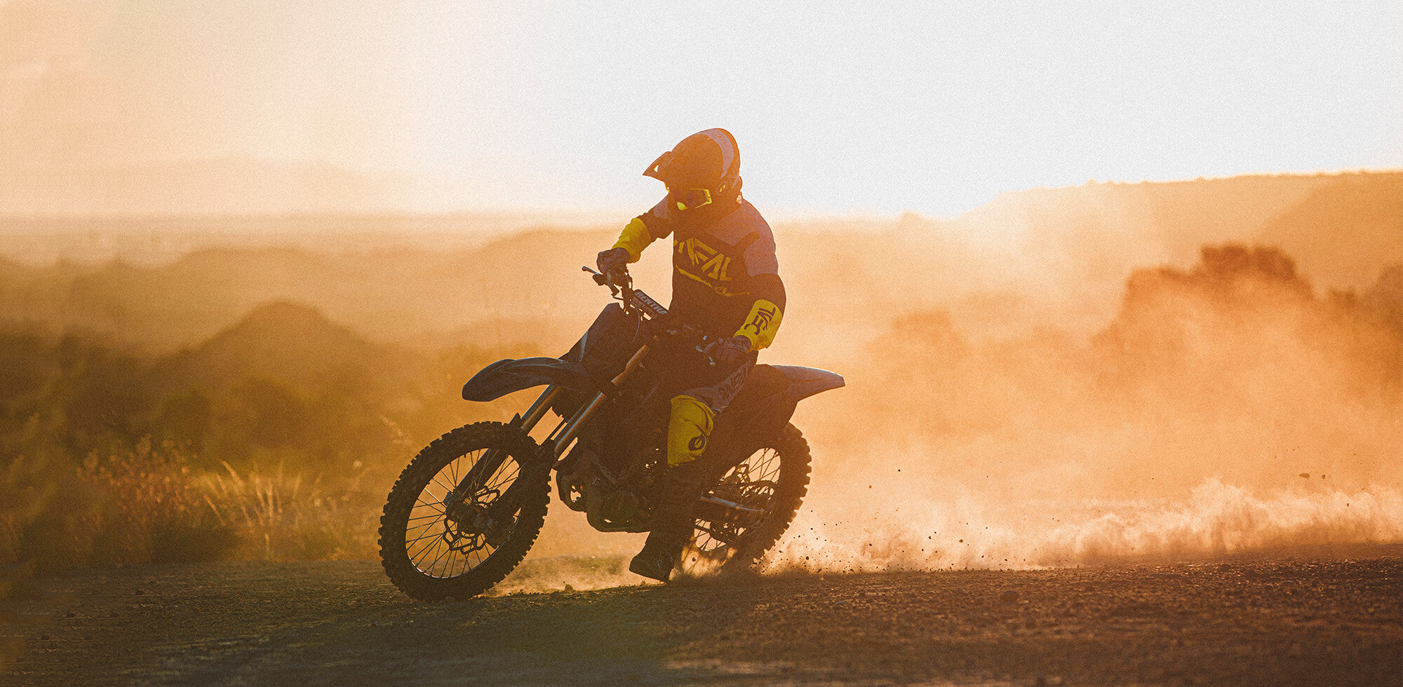 Ein Motorradfahrer in gelb-schwarzer Motorradmontur legt sich bei Sonnenuntergang in eine Kurve auf einer unbefestigten Straße und wirbelt dabei Staub auf. Das dunstige, goldene Licht erzeugt einen dramatischen Effekt, im Hintergrund sind zerklüftetes Gelände und Hügel zu sehen.