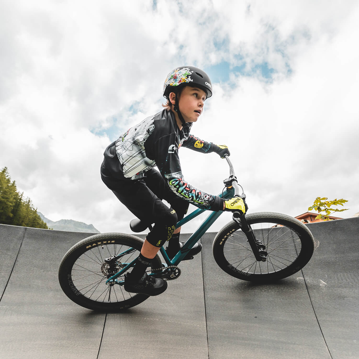 Ein junger Mensch mit Helm und bunter Sportbekleidung fährt mit einem BMX-Rad eine geschwungene Rampe entlang. Der Himmel ist bewölkt, im Hintergrund sind Bäume und ein Gebäude zu sehen. Der Fahrer wirkt konzentriert und lehnt sich mit seinem Rad in die Kurve.