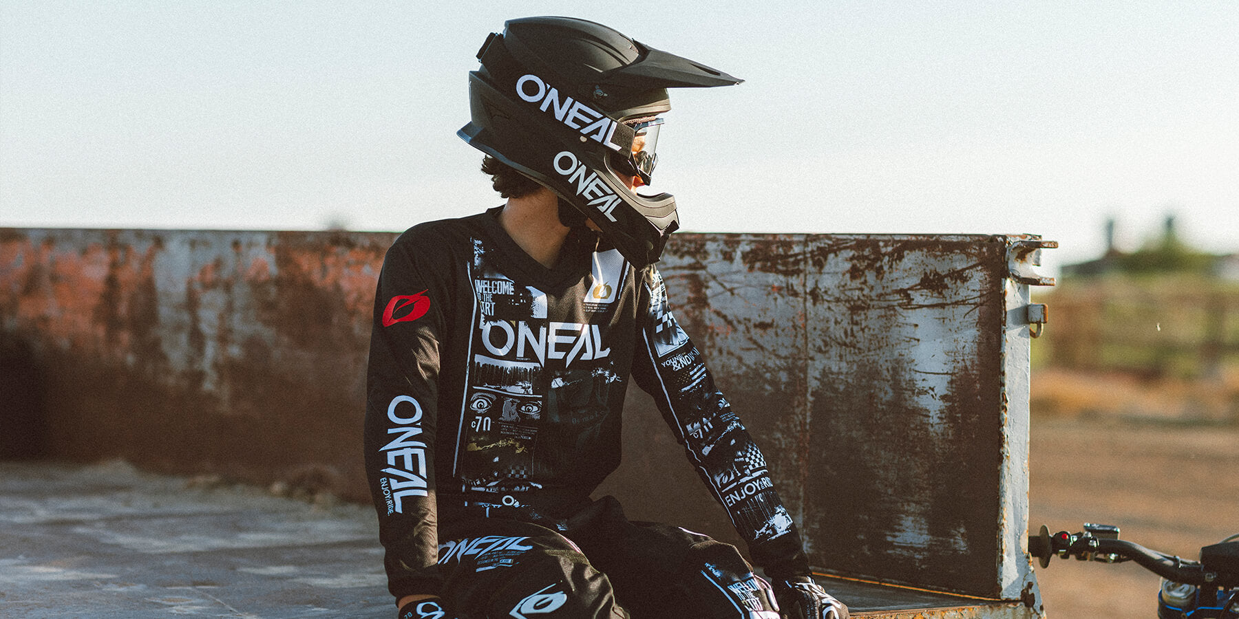 Ein Motocrossfahrer in komplett schwarz-weißer O'Neal-Ausrüstung, einschließlich Helm und Trikot, sitzt auf einer verrosteten Metallplattform. Der Fahrer blickt nach rechts, hinter ihm sind eine Feldstrecke und ein unscharfer Hintergrund zu sehen.