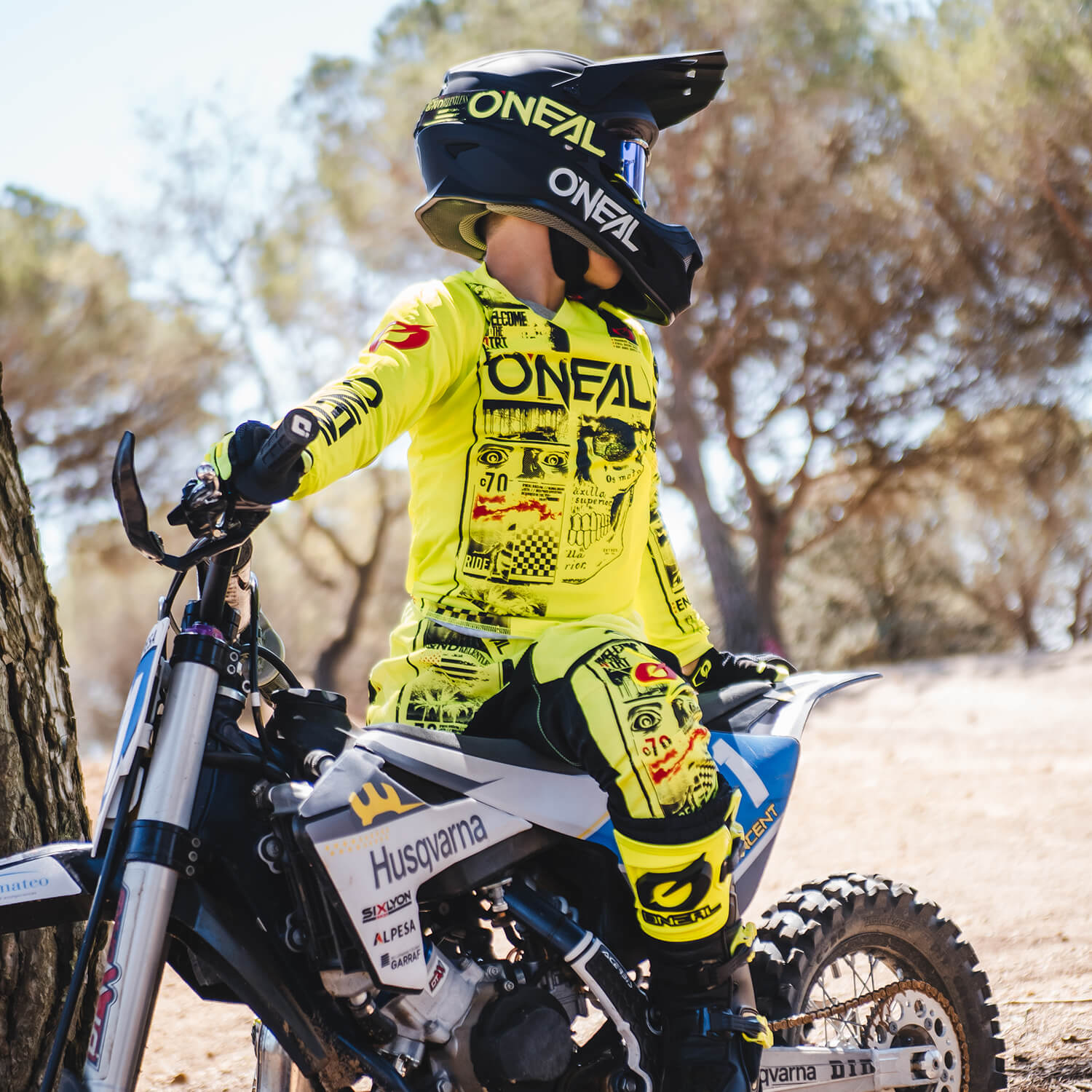 Eine Person in einem leuchtend gelb-schwarzen O'Neal-Motocross-Outfit und Helm sitzt auf einem Husqvarna-Dirtbike und blickt zur Seite. Die Szene spielt im Freien mit trockenem Boden und Bäumen im Hintergrund.
