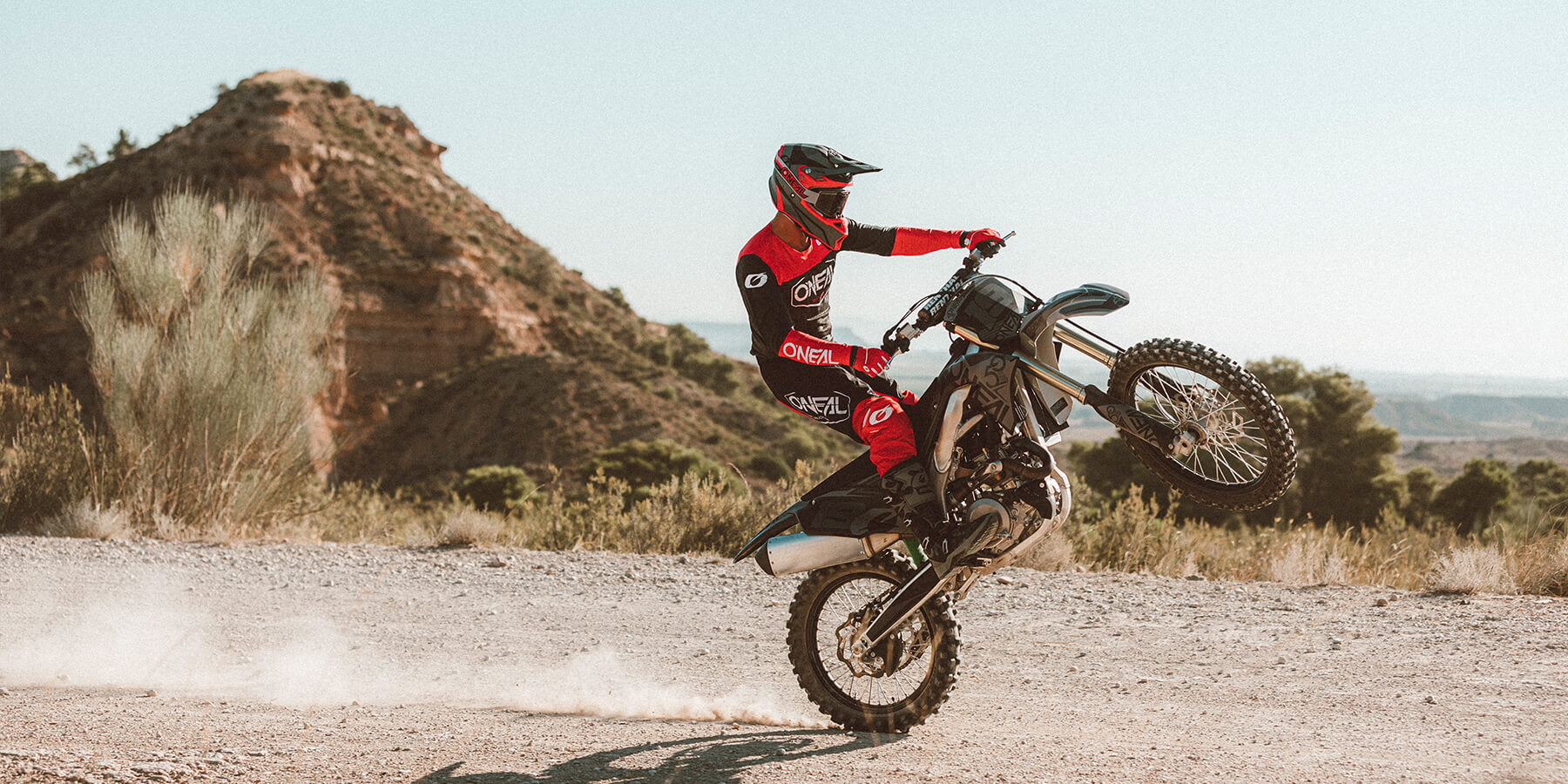Eine Person in rot-schwarzer Motocross-Ausrüstung macht mit einem Dirtbike in einer zerklüfteten, felsigen Landschaft einen Wheelie. Vom Hinterrad des Motorrads wirbelt Staub auf, im Hintergrund ist unter einem klaren Himmel eine große Felsformation zu sehen.