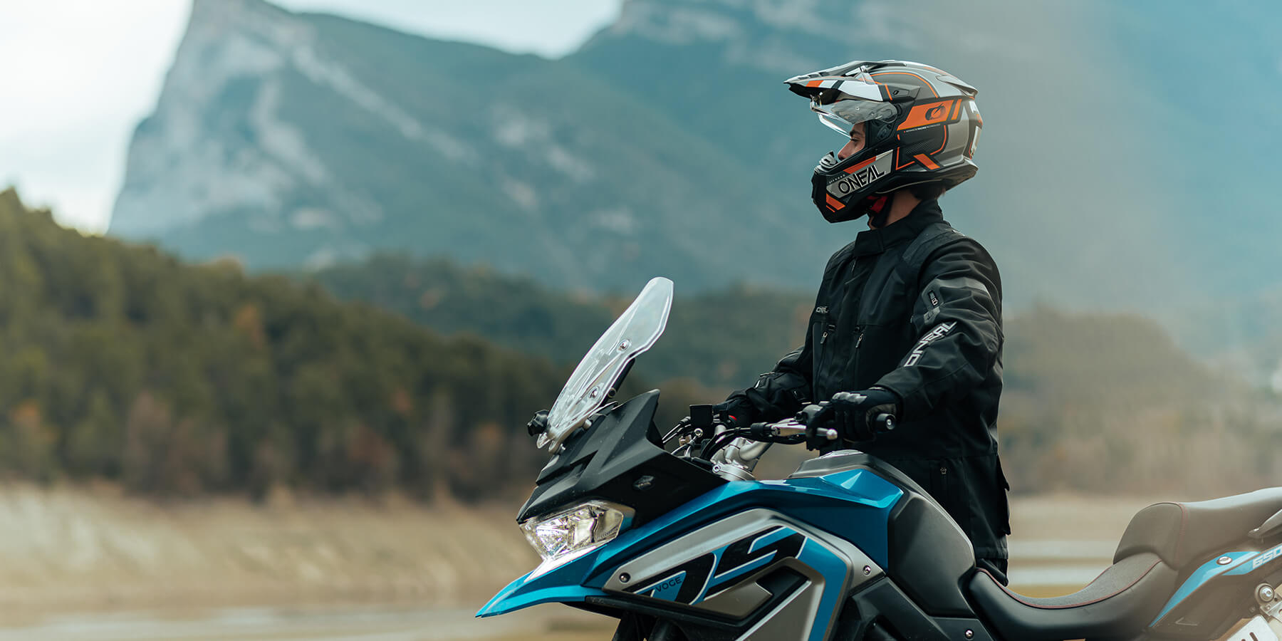 Eine Person mit schwarzer Jacke, Handschuhen und Helm fährt auf einem blau-schwarzen Motorrad auf einer Panoramastraße mit Bergen und Grün im Hintergrund. Die Person blickt geradeaus und navigiert durch die ruhige Landschaft.