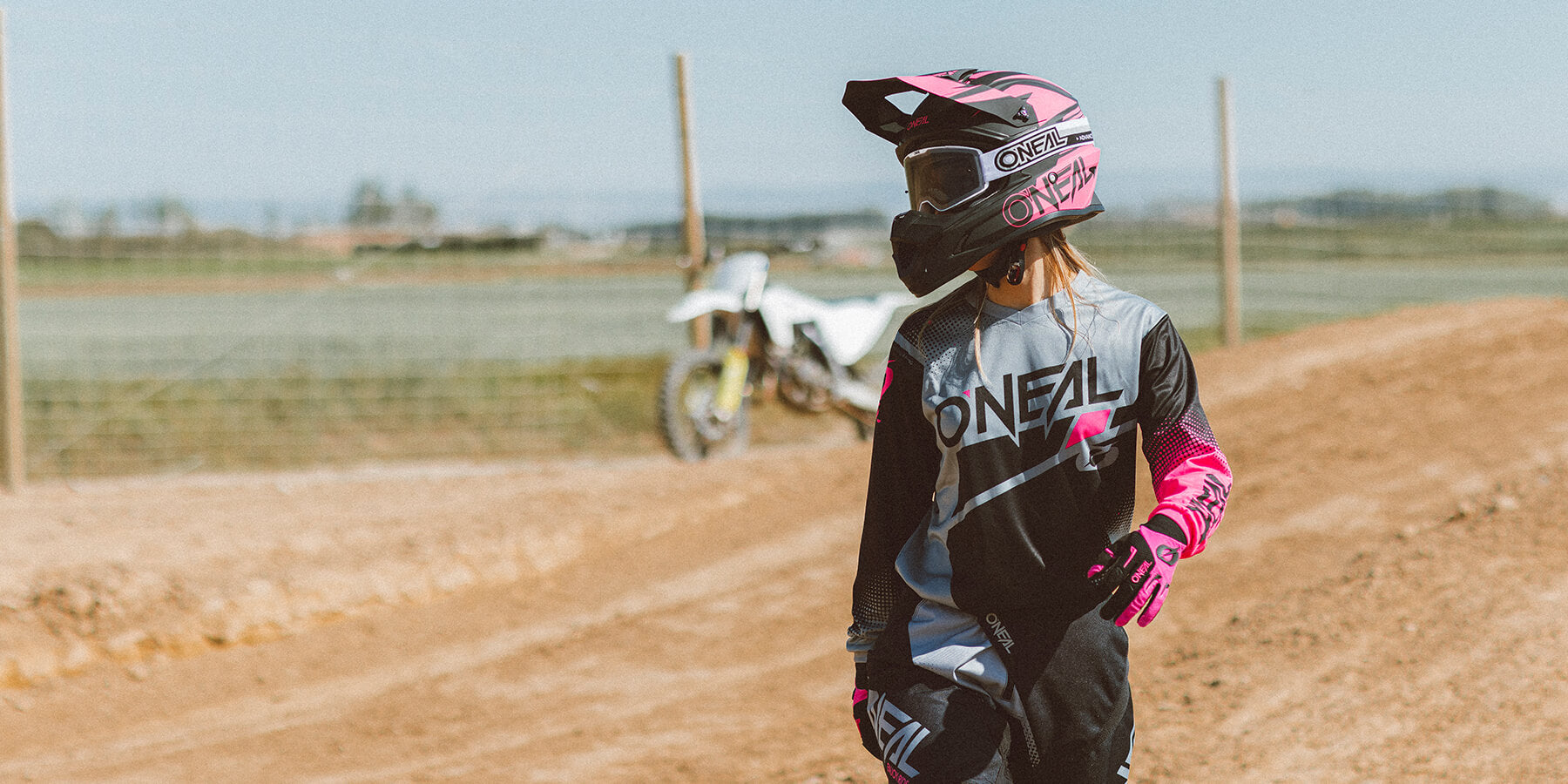 Eine Person in einem schwarz-grau-rosa Motocrossanzug und Helm steht auf einer unbefestigten Strecke. Im Hintergrund ist an einem klaren, sonnigen Tag neben einem Maschendrahtzaun ein weißes Motocross-Motorrad geparkt.