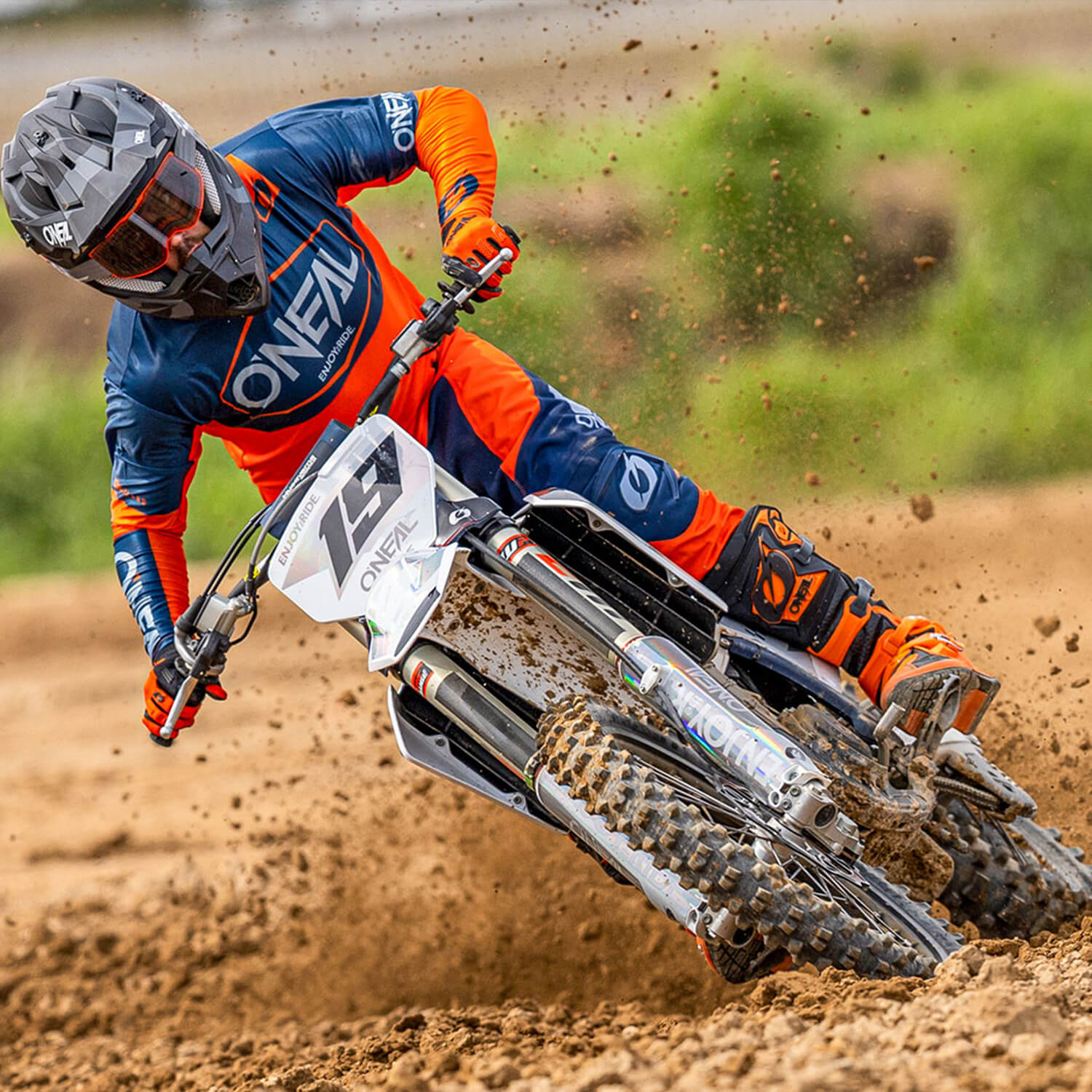 Ein Motocrossfahrer in orange-blauem Outfit, Schutzausrüstung und Helm fährt auf einer unbefestigten Strecke eine scharfe Kurve. Der Vorderreifen des Motorrads ist leicht angehoben und wirbelt Staub auf, was die Geschwindigkeit und Intensität des Sports veranschaulicht.