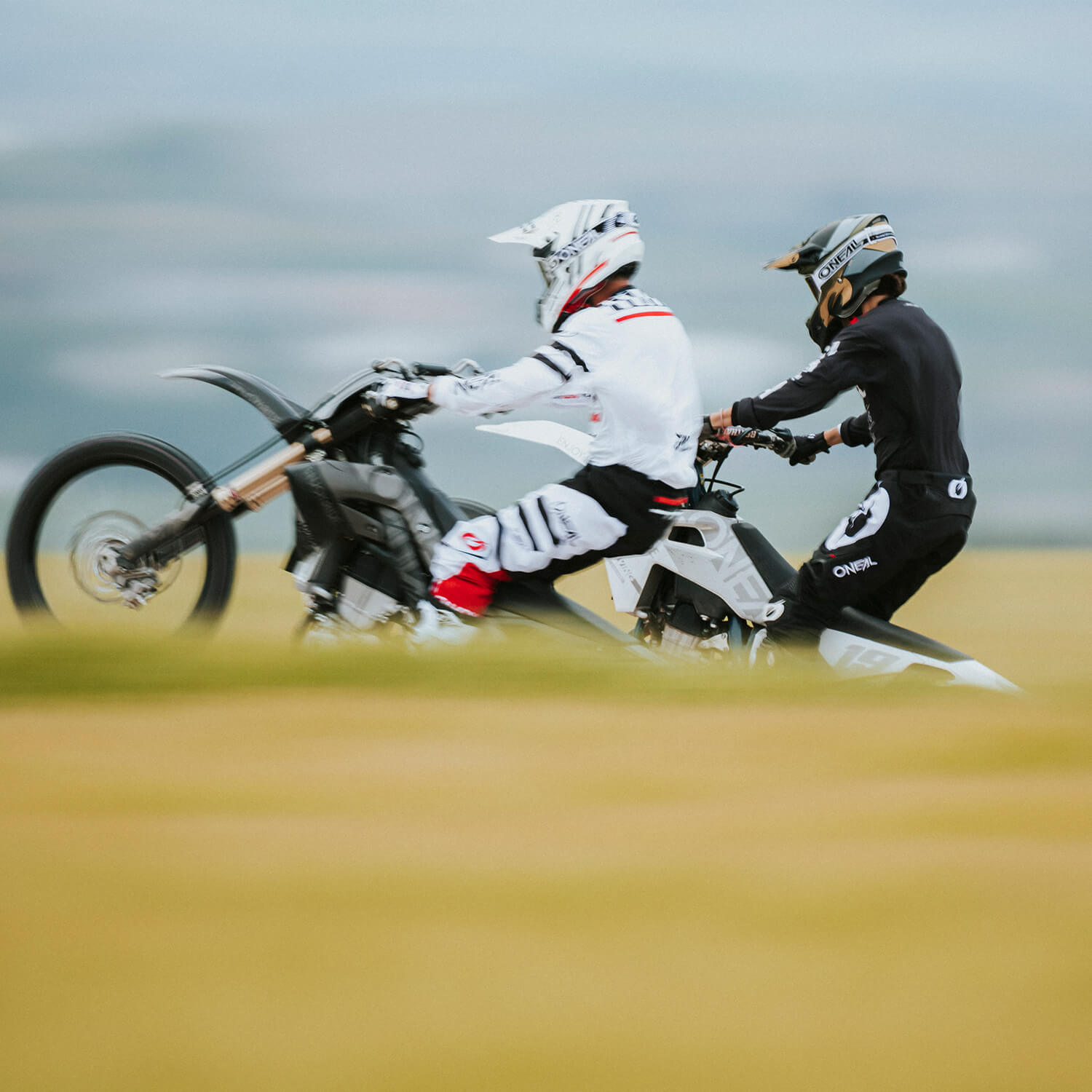 Zwei Motorradfahrer, einer in weißer Kleidung, der einen Wheelie macht, und der andere in schwarzer Kleidung, der parallel fährt, sind in Bewegung durch eine verschwommene Landschaft aufgenommen. Die Szene lässt auf eine Outdoor-Aktivität schließen, möglicherweise Motocross oder ein Offroad-Rennen.