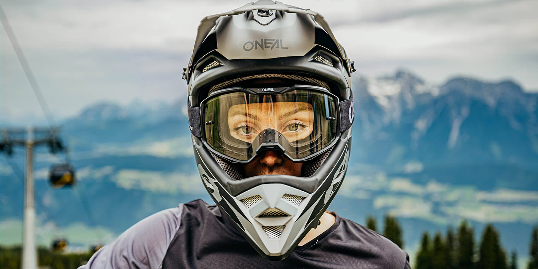 Eine Nahaufnahme einer Person mit Integralhelm und Schutzbrille, die im Freien steht, im Hintergrund eine Berglandschaft. Die Person blickt direkt in die Kamera und auf dem Helm ist die Marke „Oneal“ zu sehen.