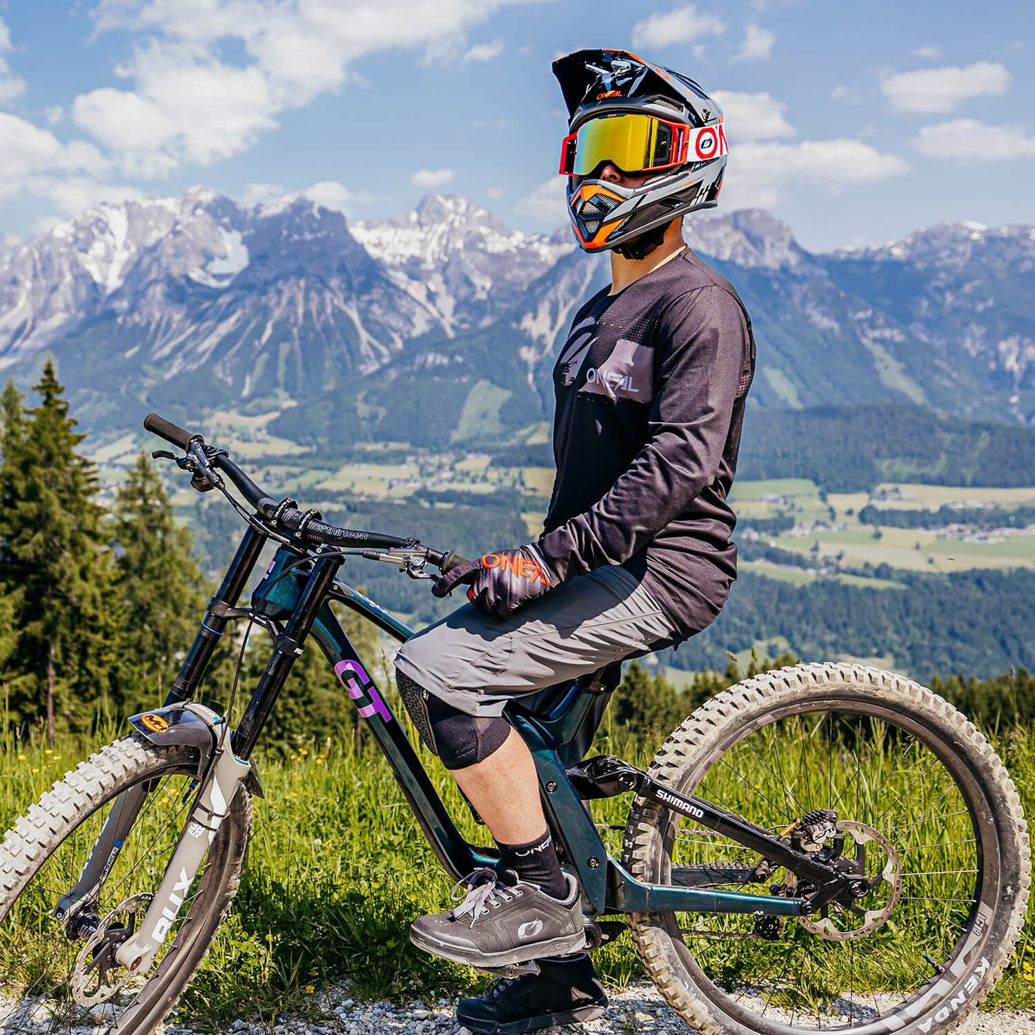 Ein Mountainbiker in voller Montur, einschließlich Helm, Schutzbrille und Handschuhen, sitzt auf einem schwarzen GT-Bike vor einer Berglandschaft mit klarem Himmel im Hintergrund. Der Fahrer trägt ein schwarzes Langarmshirt und graue Knieschützer. Unter ihm sind grüne Felder und Wälder zu sehen.