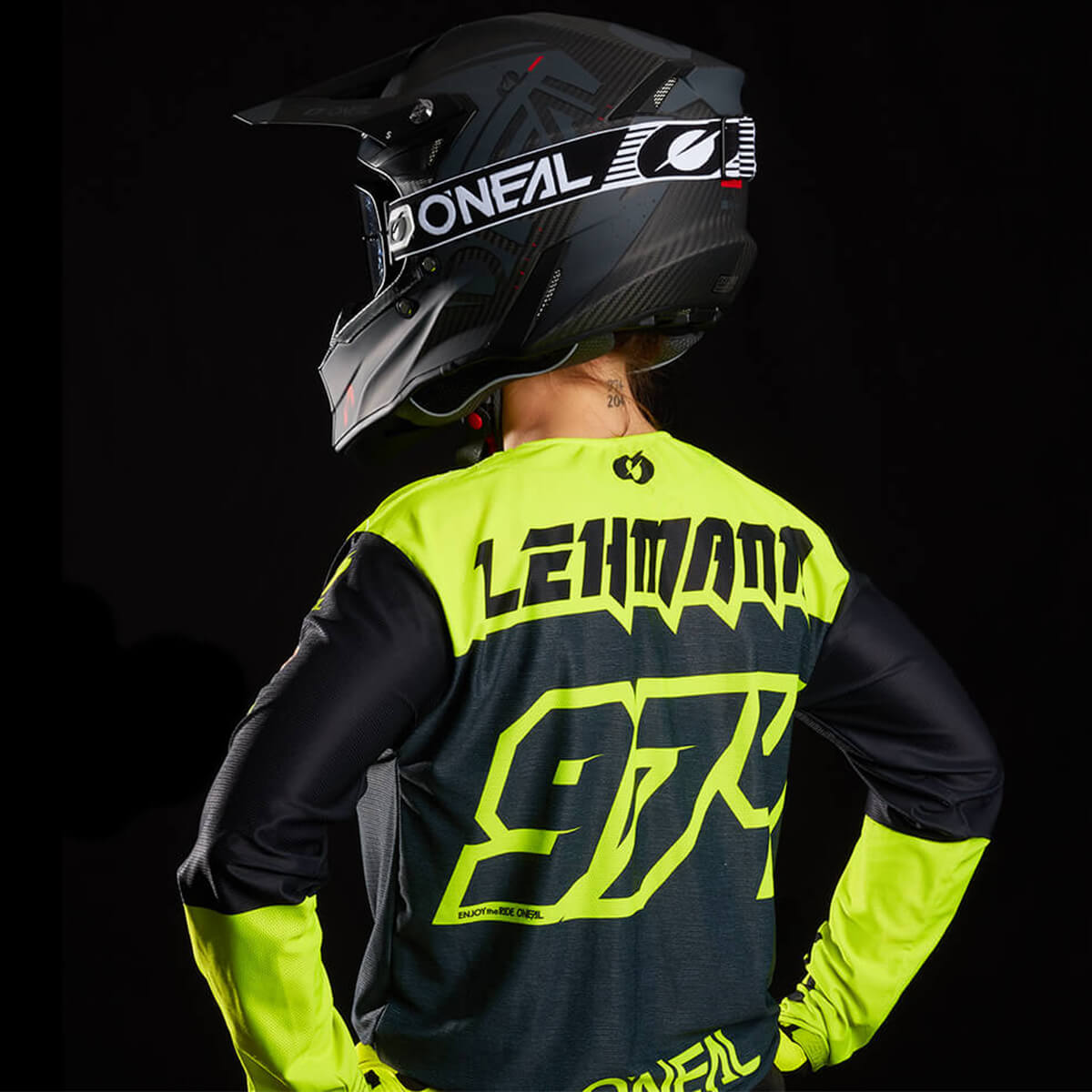 Eine Person, die ein schwarz-neongelbes Motocross-Trikot mit der Aufschrift „Leimann 974“ auf dem Rücken und einen schwarzen Integralhelm mit der Aufschrift „ONEAL“ trägt, steht mit den Händen in den Hüften vor einem dunklen Hintergrund.