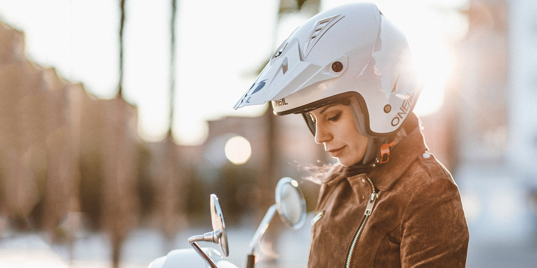 Eine Person mit weißem Motorradhelm und brauner Jacke steht neben einem Motorroller. Der Kopf der Person ist leicht nach unten geneigt und blickt in den Spiegel des Motorrollers. Im Hintergrund scheint helles Sonnenlicht.
