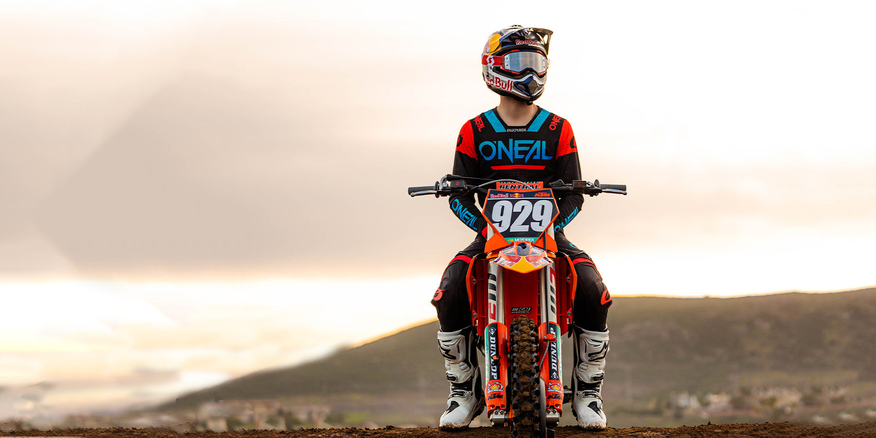 Ein Motocrossfahrer mit Helm, Schutzbrille und leuchtend rot-blauem Outfit mit der Nummer 929 auf dem Motorrad sitzt auf einem orangefarbenen Dirtbike. Im Hintergrund ist eine Berglandschaft unter einem bewölkten Himmel zu sehen.