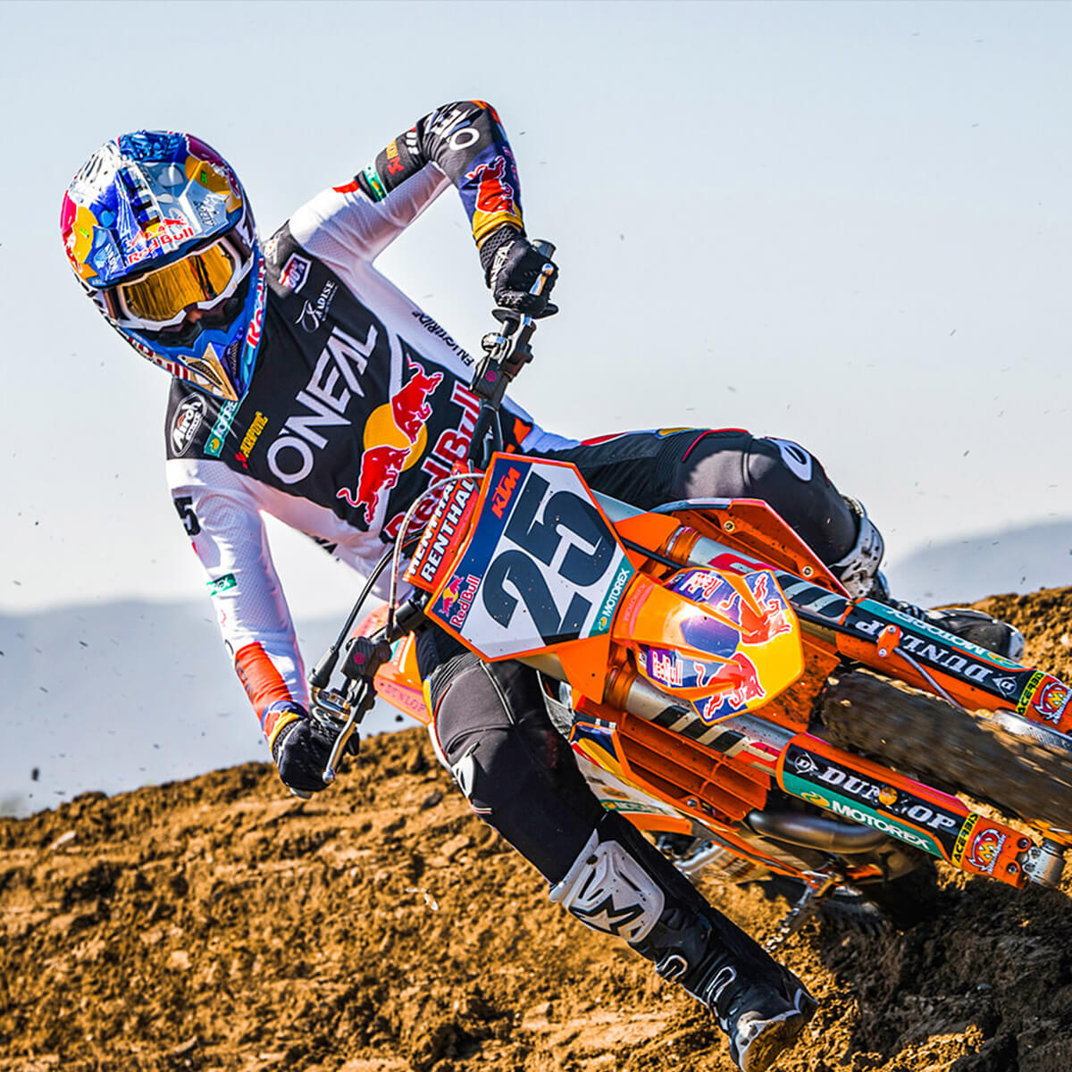 Ein Motocrossfahrer mit buntem Helm, Schutzbrille und Marken-Rennausrüstung lehnt sich auf einem orange-schwarzen Motorrad in eine Dirt-Kurve. Auf dem Motorrad ist die Zahl „25“ zu sehen. Im Hintergrund ist ein klarer Himmel und eine staubige Piste zu sehen.