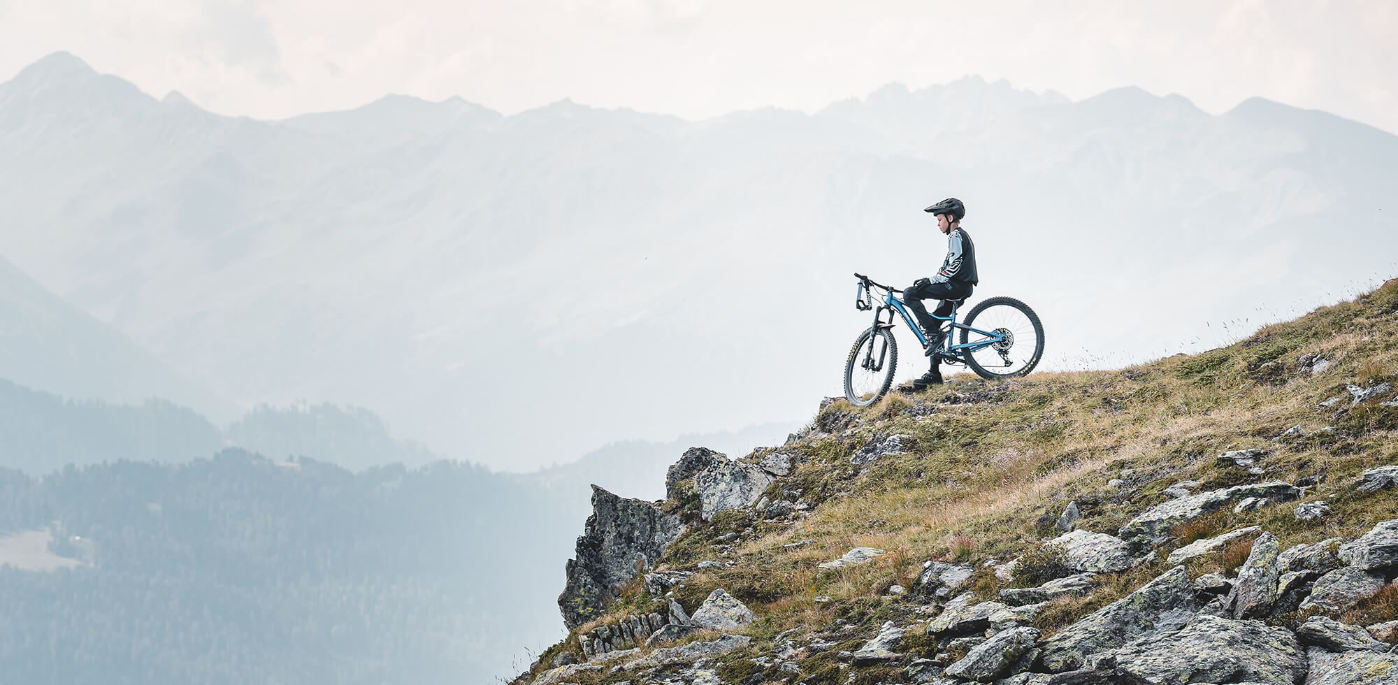 Ein Mountainbiker hält am Rand einer Felswand inne, das Fahrrad neben sich, und überblickt eine weite Landschaft mit fernen Berggipfeln unter einem bewölkten Himmel. Die Szene strahlt Ruhe und ein Gefühl von Abenteuer aus.