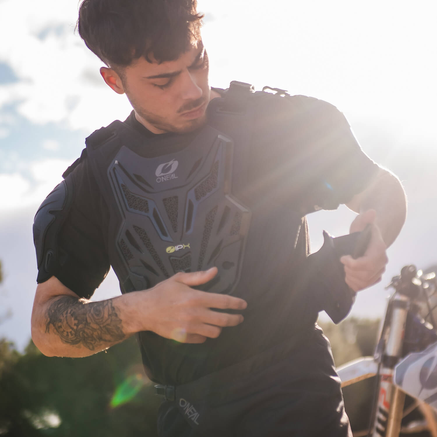 Eine Person steht im Freien im Sonnenlicht und trägt einen schwarzen Brustschutz von O'Neal und passt die Riemen an. Im Hintergrund ist teilweise ein Motocross-Motorrad zu sehen. Die Person hat kurze dunkle Haare und sichtbare Tätowierungen auf dem linken Arm.