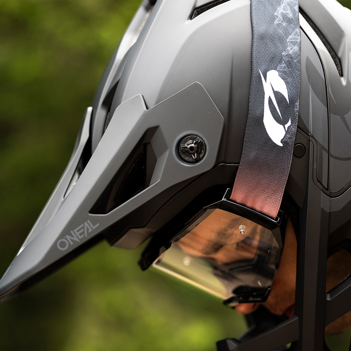 Nahaufnahme einer Person mit einem mattgrauen O’Neal-Fahrradhelm mit Visier und transparenter Schutzbrille vor einem verschwommenen grünen Außenhintergrund. Der Helmriemen ist mit einem weißen Logo versehen.