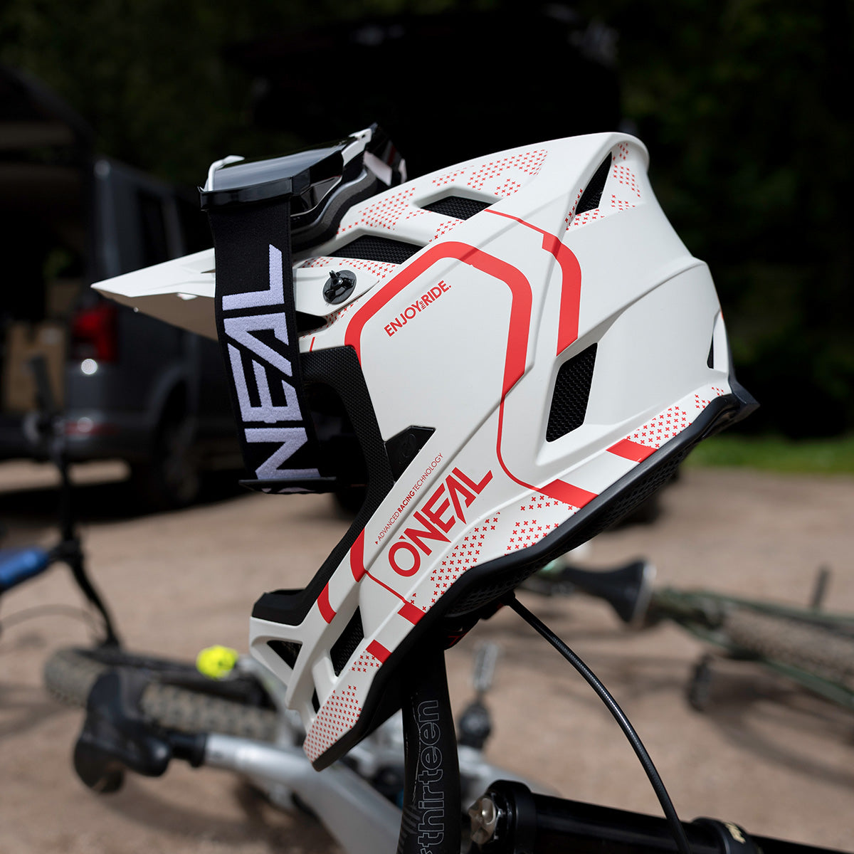 Eine Nahaufnahme eines weiß-roten O’NEAL-Integralhelms für Mountainbikes mit schwarzem Riemen, der auf einem Fahrradsitz ruht, mit verschwommenen Fahrrädern und einem Fahrzeug im Hintergrund.
