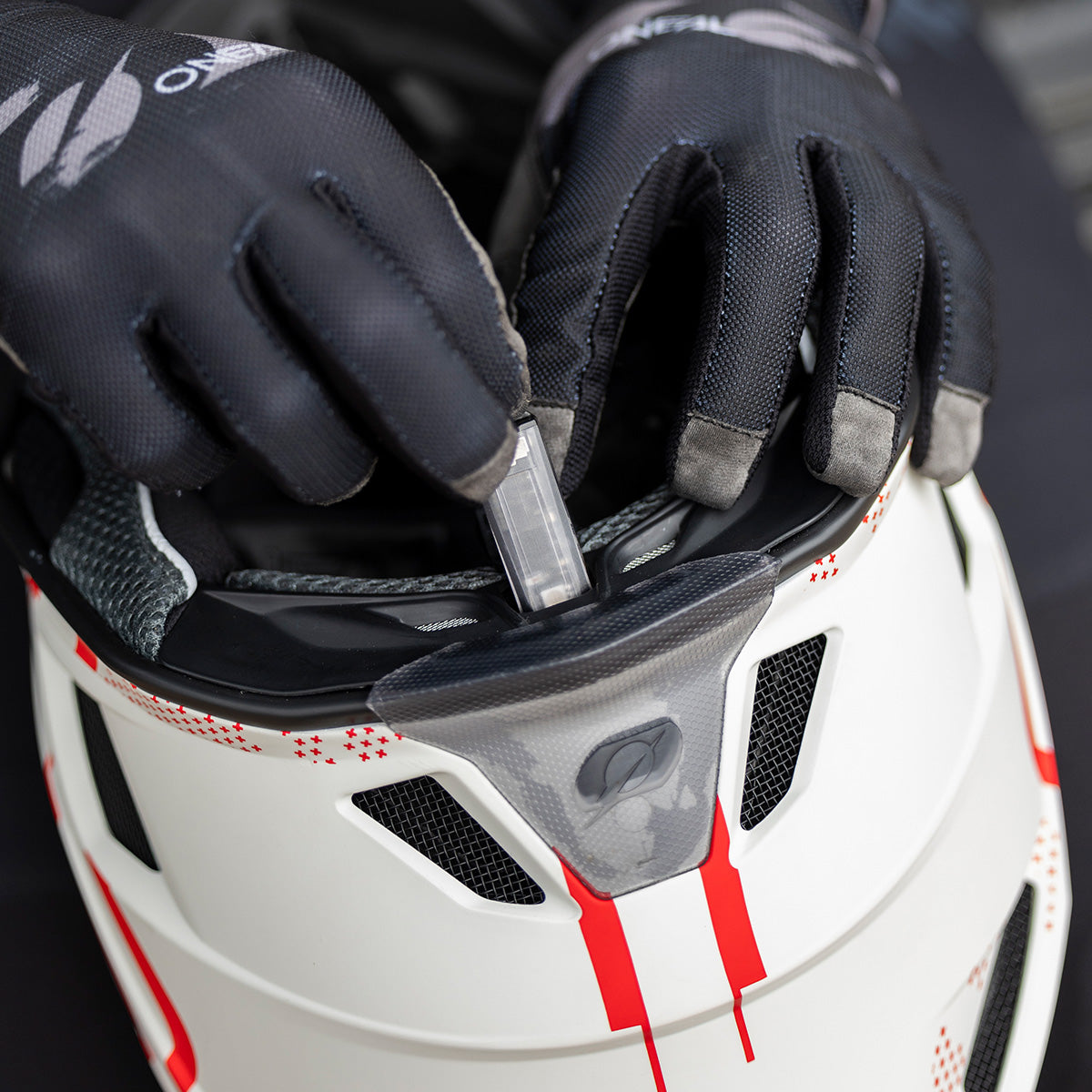 Eine Person mit schwarzen Handschuhen passt einen kleinen rechteckigen Gegenstand an oder steckt ihn in den Kinnbereich eines weiß-roten Motorradhelms.