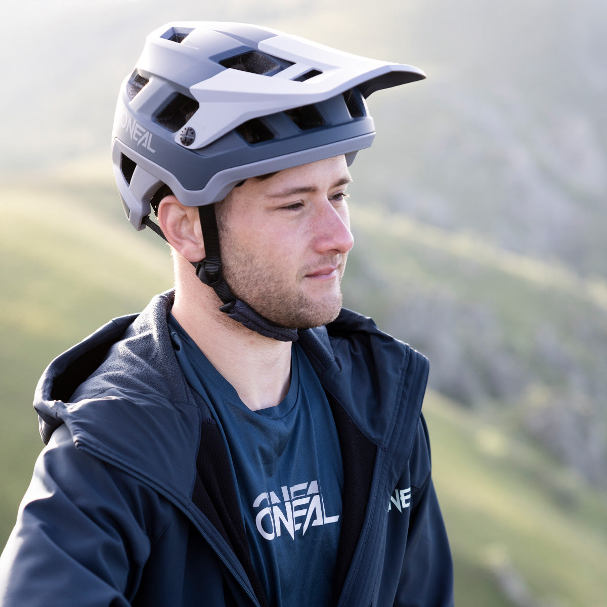 Ein Mann in einer blauen O'Neal-Jacke und einem Helm steht im Freien in einer bergigen Gegend und blickt mit neutralem Gesichtsausdruck zur Seite.