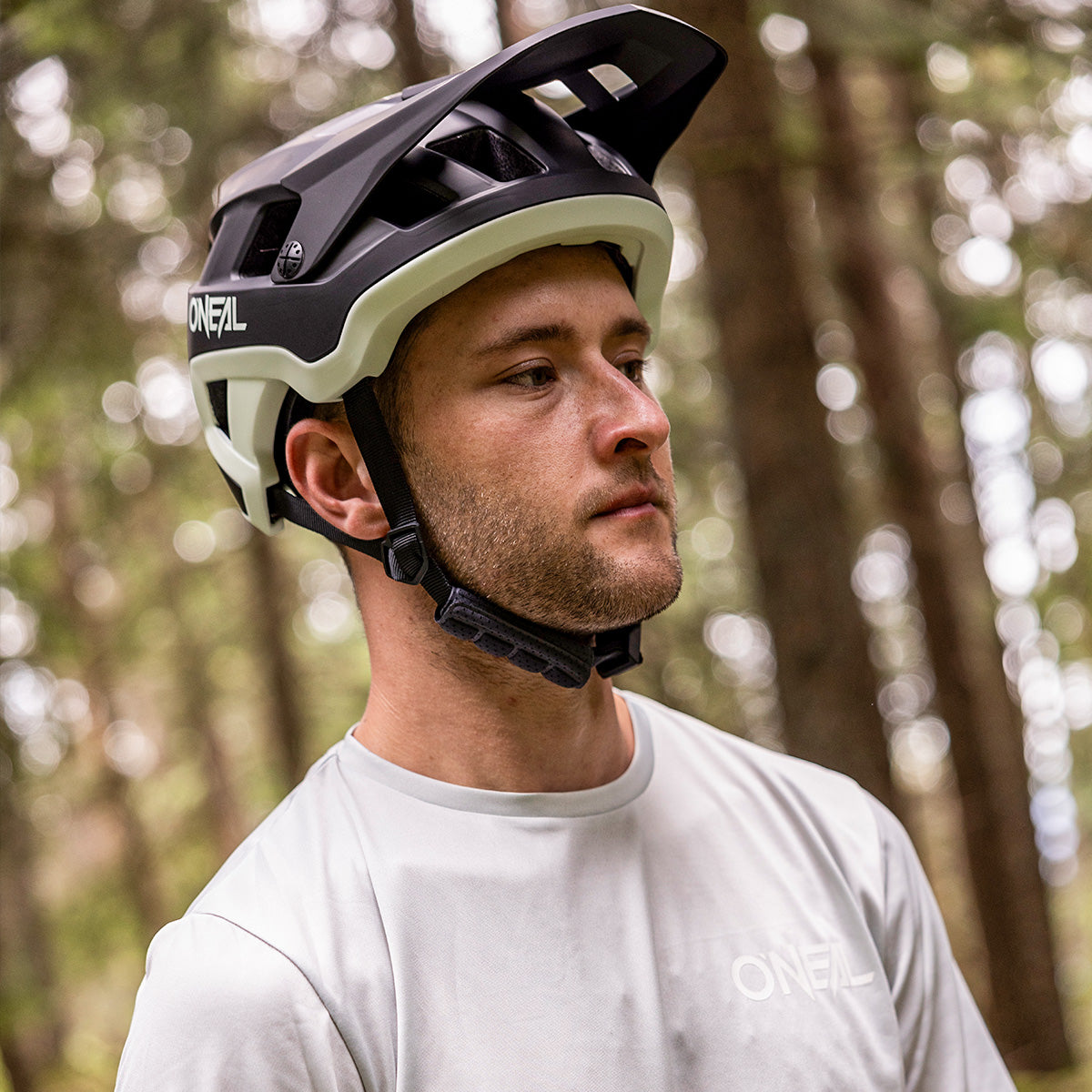 Ein Mann mit einem schwarz-weißen O'Neal-Fahrradhelm und einem hellen O'Neal-Shirt steht in einem Wald und blickt nachdenklich in die Ferne. Im Hintergrund sind hohe Bäume mit grünem Laub verschwommen.
