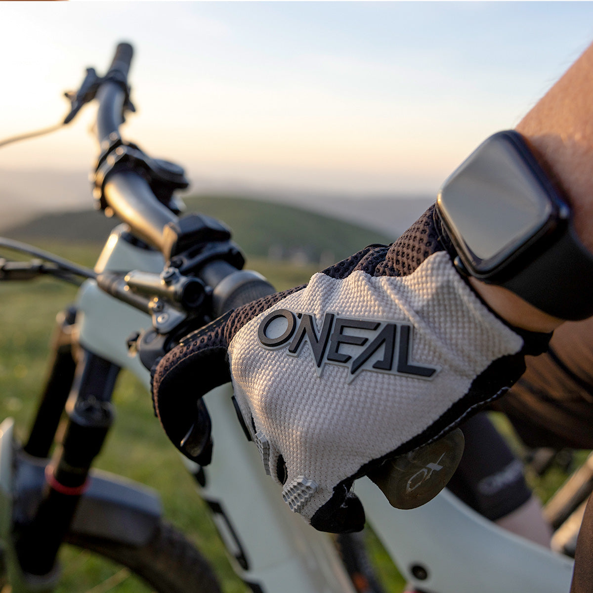 Nahaufnahme einer Person mit O'NEAL-Handschuhen und Smartwatch, die im Freien den Lenker eines Mountainbikes umklammert, mit einer verschwommenen Landschaft im Hintergrund bei Sonnenuntergang.