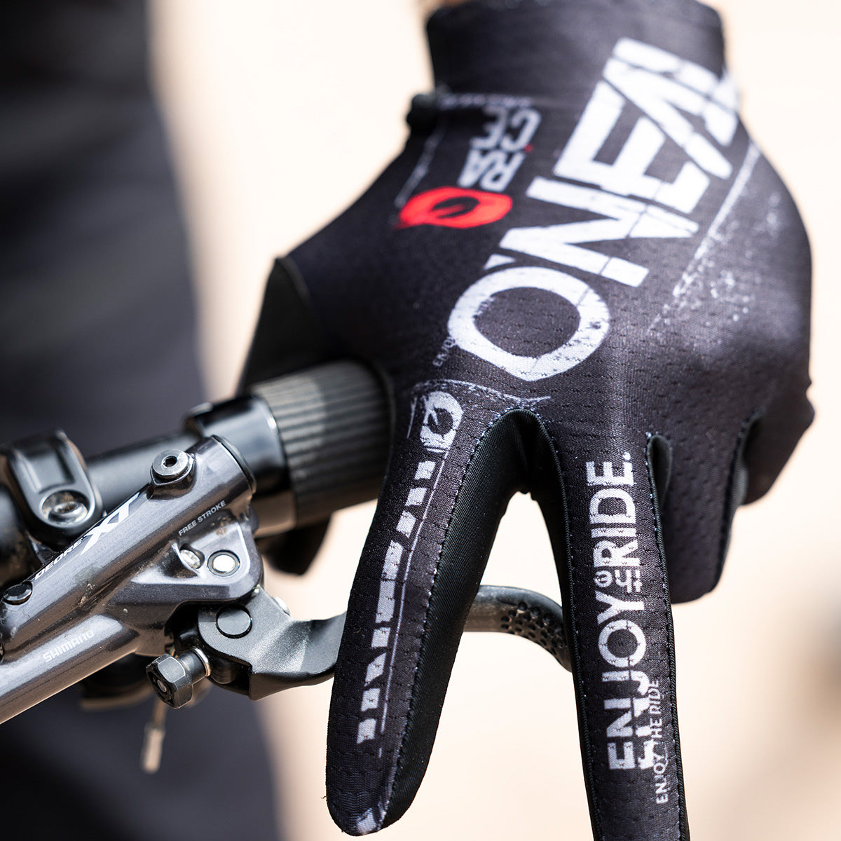 Eine Nahaufnahme einer Hand mit einem schwarzen O'NEAL-Fahrradhandschuh, die einen Fahrradlenker umklammert. Auf dem Handschuh sind in weißer Schrift die Sätze „ENJOY THE RIDE“ und „O'NEAL“ zu sehen.