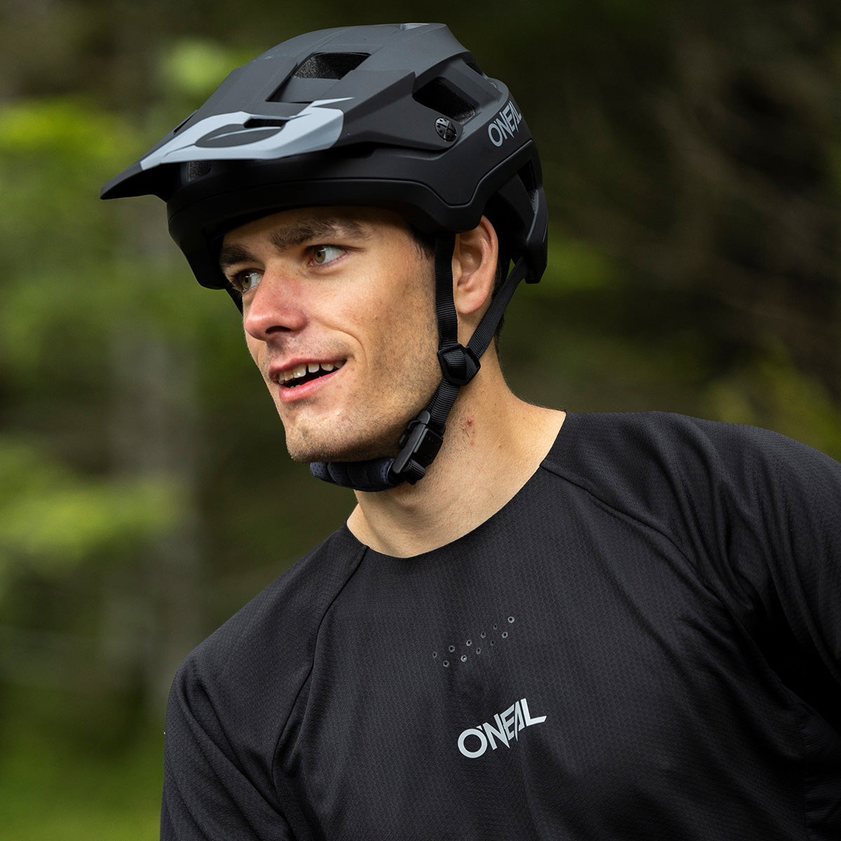 Ein Mann mit einem schwarzen Mountainbike-Helm und einem schwarzen O’Neal-Trikot blickt nach draußen zur Seite, vor dem Hintergrund eines verschwommenen grünen Waldes.