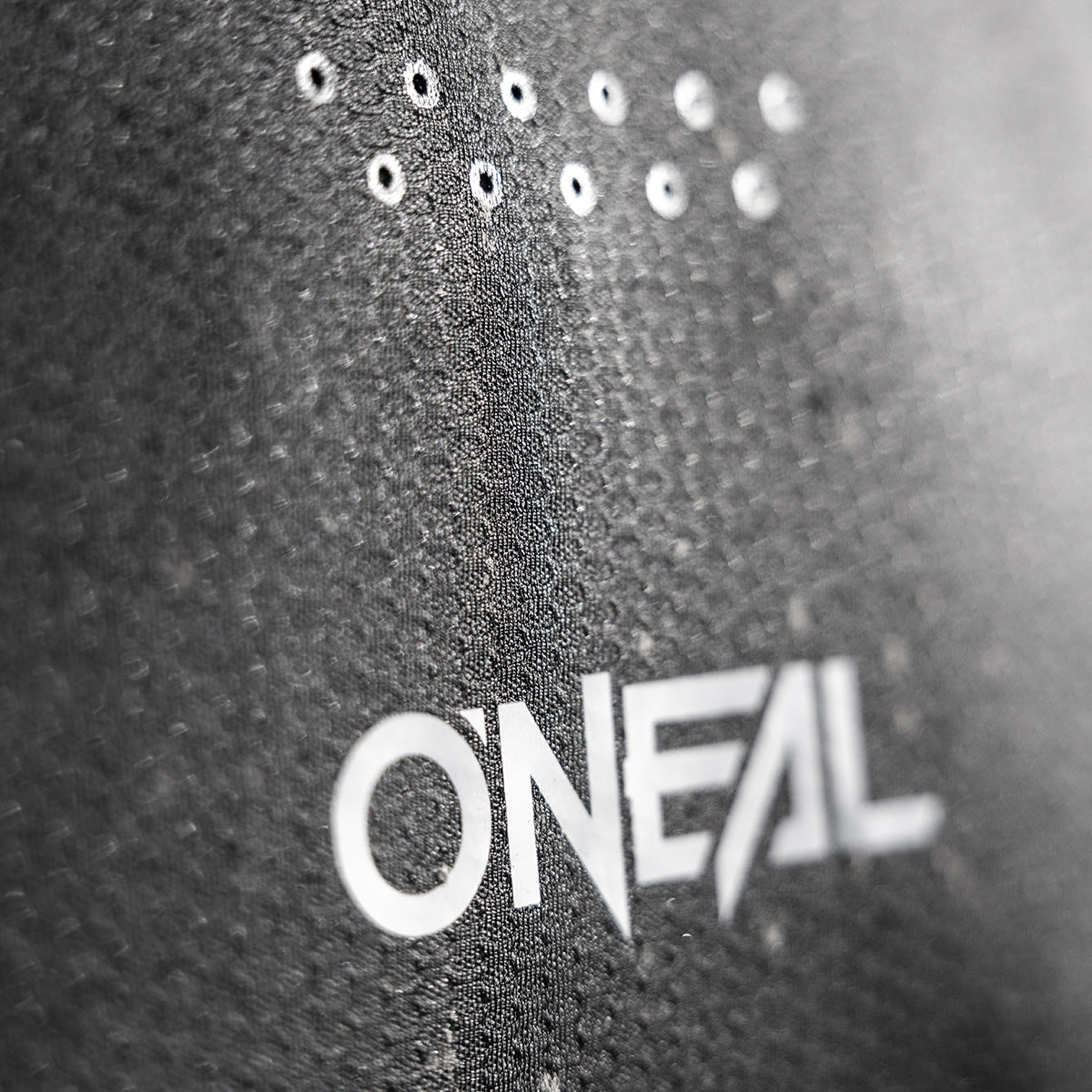 Nahaufnahme eines strukturierten schwarzen Stoffes mit dem weißen „ONEAL“-Logo und zwei Reihen kleiner runder Belüftungslöcher darüber.