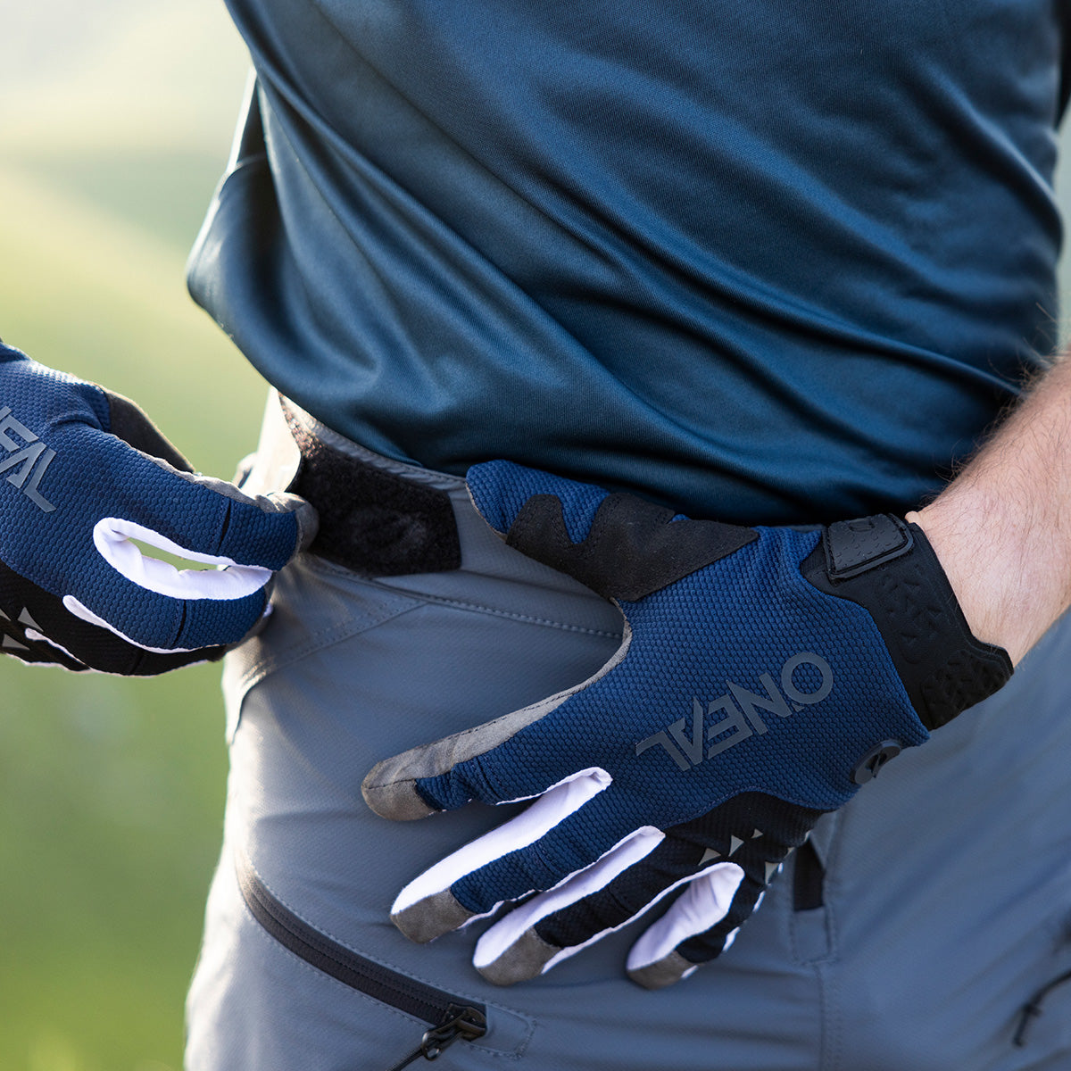 Eine Person mit blauen Fahrradhandschuhen und einem türkisfarbenen Hemd rückt den Bund einer grauen Outdoorhose zurecht und schließt den Klettverschluss. Auf den Handschuhen ist der Markenname „ONEAL“ zu sehen.
