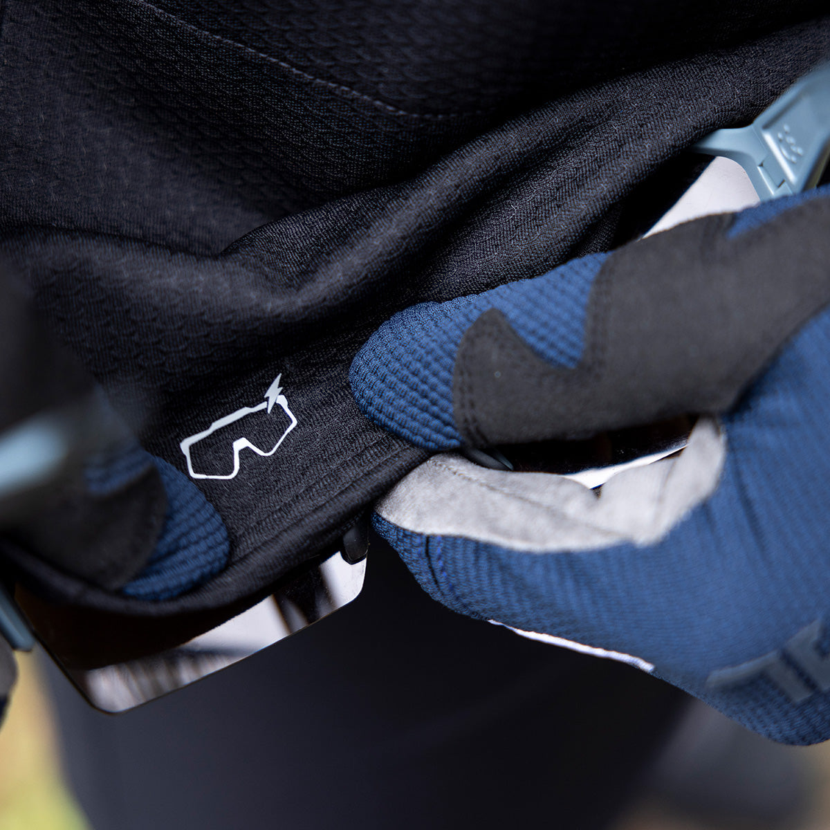 Eine Nahaufnahme behandschuhter Hände, die ein Stück schwarzen Stoff mit einem kleinen weißen Symbol einer Schutzbrille und einem Blitz halten. Die Handschuhe sind blau und grau und bestehen aus strukturiertem Material.