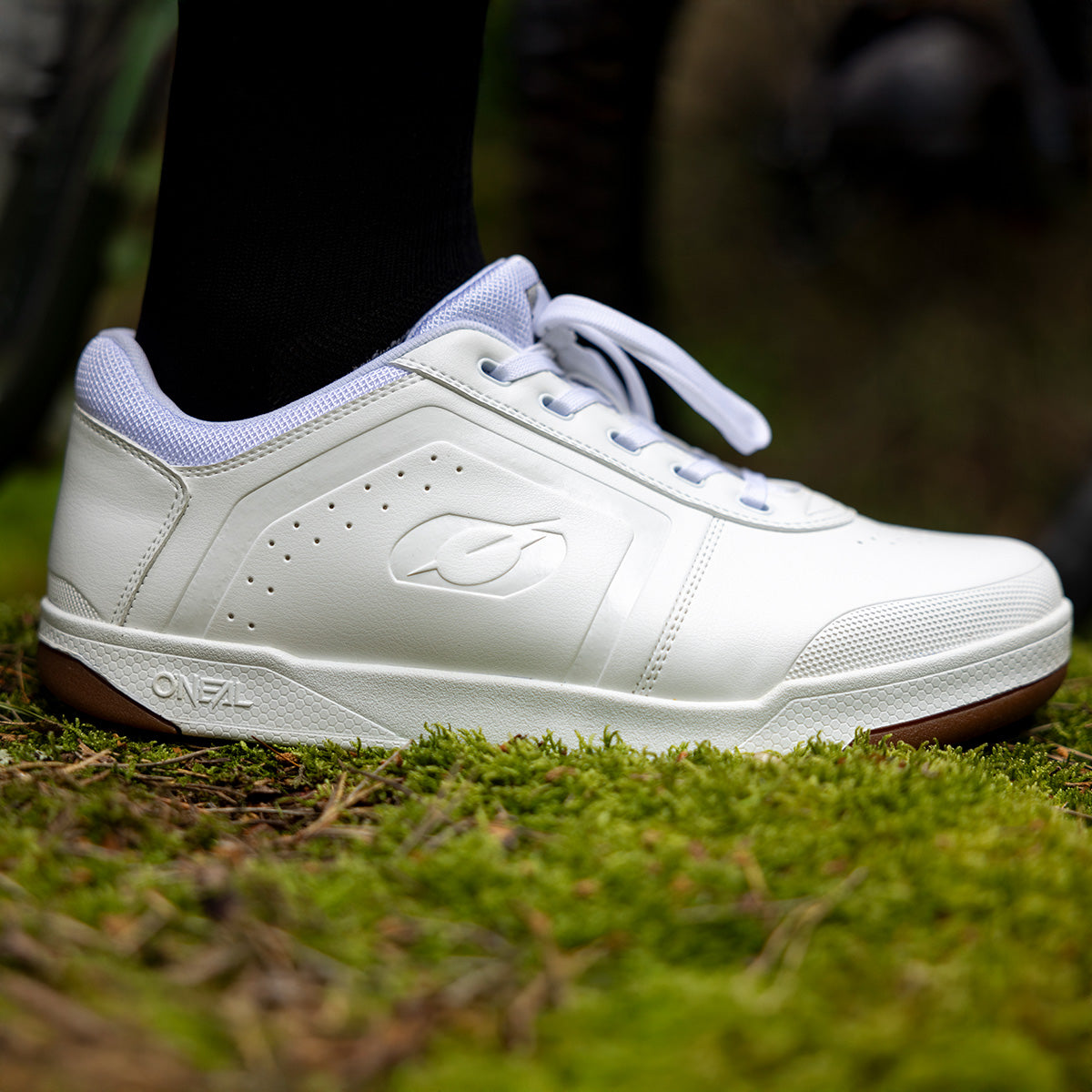 Nahaufnahme eines weißen Sportschuhs mit weißen Schnürsenkeln, der auf grünem Moosboden steht. Der Schuh hat ein dezentes Logo an der Seite und eine braune Sohle.