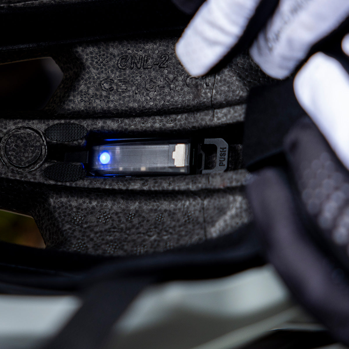 Nahaufnahme der Innenseite eines schwarzen Fahrradhelms. Man sieht ein kleines Gerät mit einem leuchtend blauen Licht und der Aufschrift „PUSH“ sowie eine Person mit schwarz-weißen Handschuhen, die es einstellt oder überprüft.