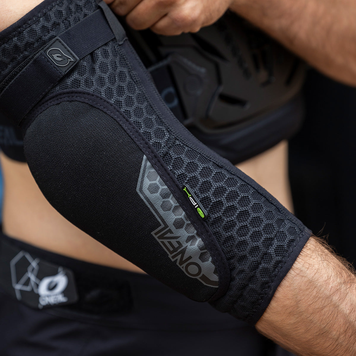 Eine Person trägt einen schwarzen, gepolsterten Ellbogenschutz mit Wabenmuster und der Aufschrift „ONEAL“ am Arm, wobei ein Teil ihres Oberkörpers und ihrer Hand sichtbar ist, und passt ihre Schutzausrüstung an.