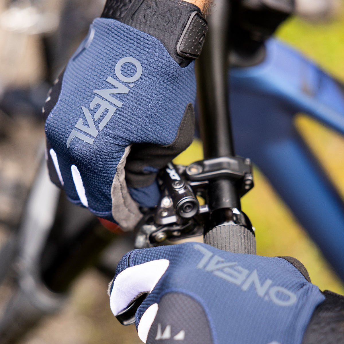 Nahaufnahme von behandschuhten Händen, die den Fahrradlenker umklammern. Die Handschuhe sind blau und schwarz und tragen die Aufschrift „ONEAL“. Der Fahrradrahmen und der Bremshebel sind teilweise sichtbar.