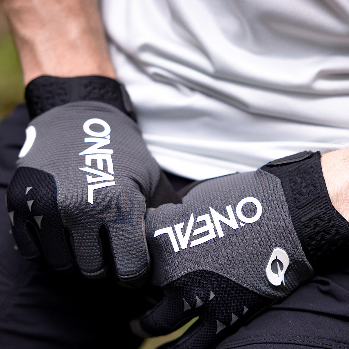 Nahaufnahme einer Person, die schwarze und graue Handschuhe der Marke O'Neal trägt, die Hände im Schoß ruhen und eine dunkle Hose und ein helles Hemd trägt.