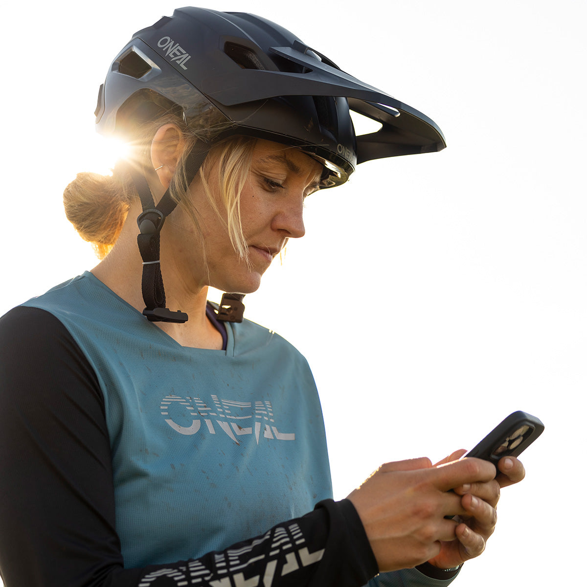 Eine Person mit einem schwarzen O'NEAL TRAILFINDER EVO Solid Quin Bundle Helm und einem blaugrünen O'NEAL Trikot blickt auf ihr Smartphone, wobei das Sonnenlicht einen warmen Schein um ihren Kopf erzeugt und den Quin Smart Safety Sensor hervorhebt.