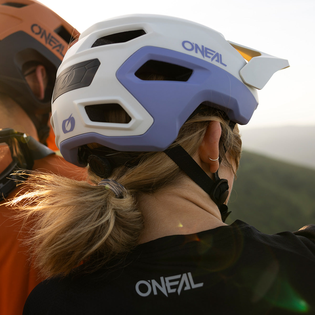 Eine Person mit hellen Haaren in einem niedrigen Pferdeschwanz trägt einen blau-weißen O'NEAL TRAILFINDER EVO Solid Quin Bundle Helm und ein schwarzes O'NEAL-Shirt. Neben ihnen steht eine Person mit einem orangefarbenen Helm im Freien mit grünen Hügeln im Hintergrund.