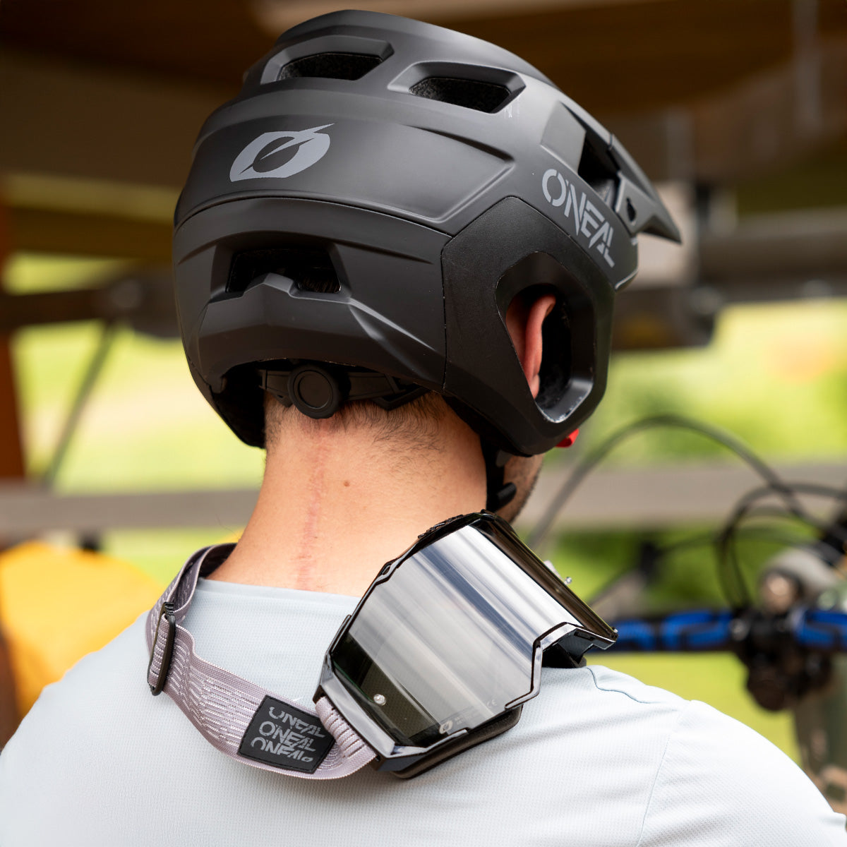 Eine Person mit einem schwarzen O’Neal-Fahrradhelm und einer grauen O’Neal-Schutzbrille um den Hals (von hinten gesehen) steht im Freien neben einem Fahrrad.
