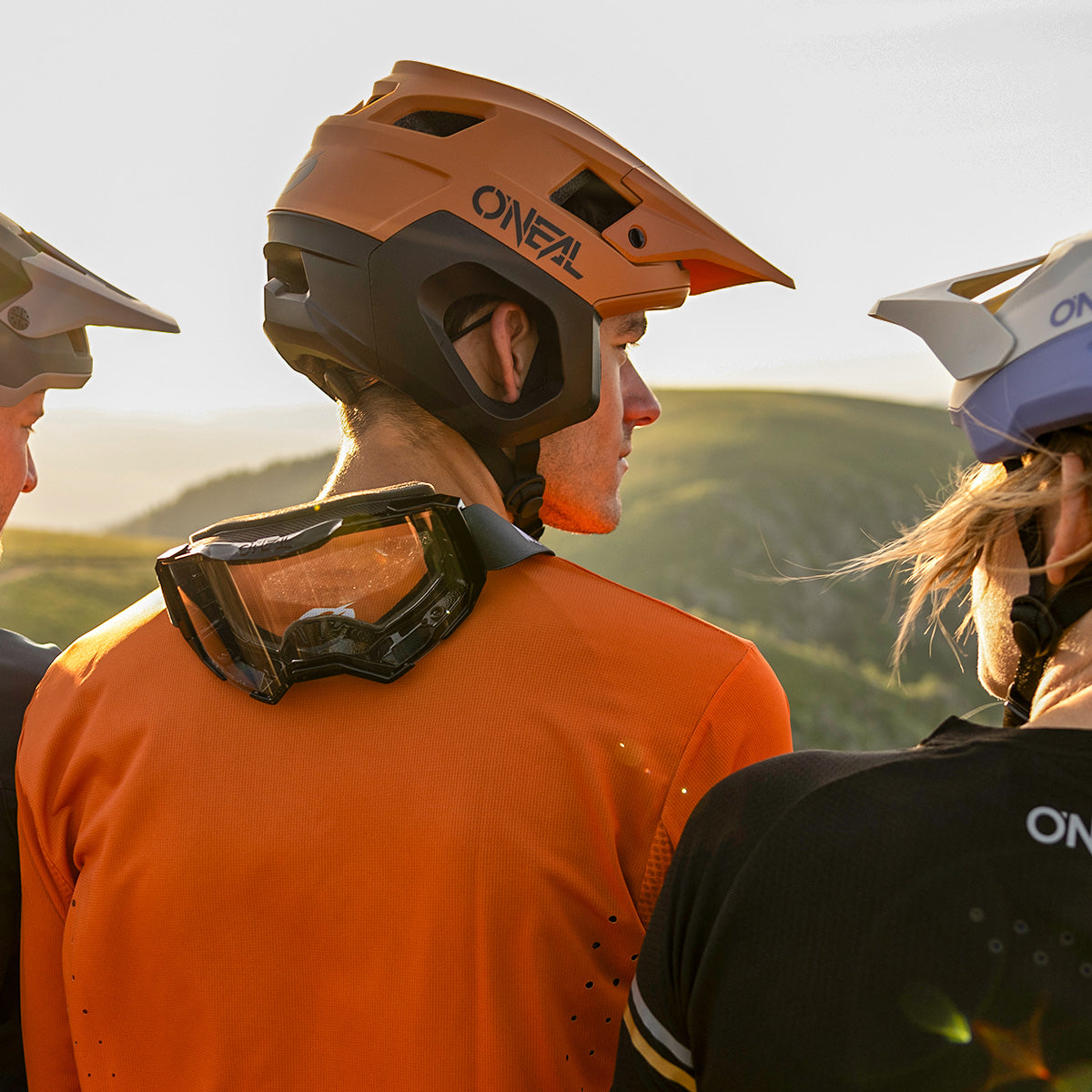 Drei Mountainbiker mit Helmen und Trikots stehen im Freien zusammen und blicken von der Kamera ab. Im Hintergrund geht die Sonne über grünen Hügeln unter. Einer der Biker trägt eine Schutzbrille im Nacken.
