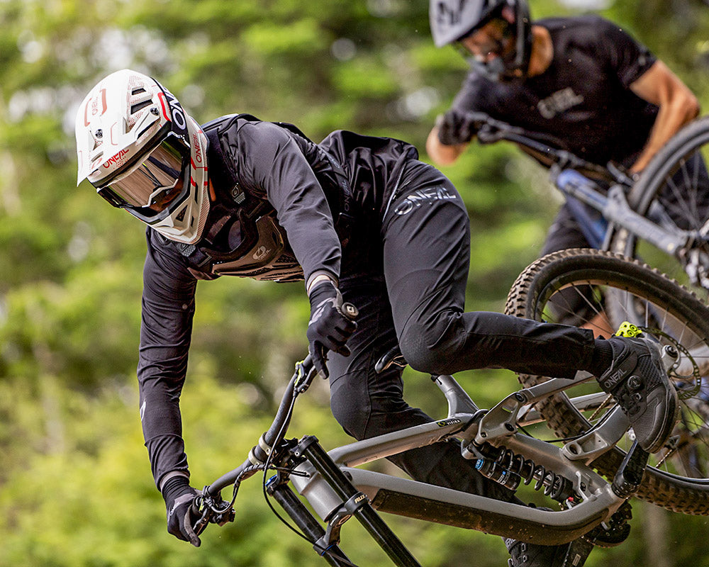 Zwei Mountainbiker mit Helmen und Schutzausrüstung fahren mit hoher Geschwindigkeit einen Waldweg bergab und legen sich mit höchster Konzentration in eine Kurve.