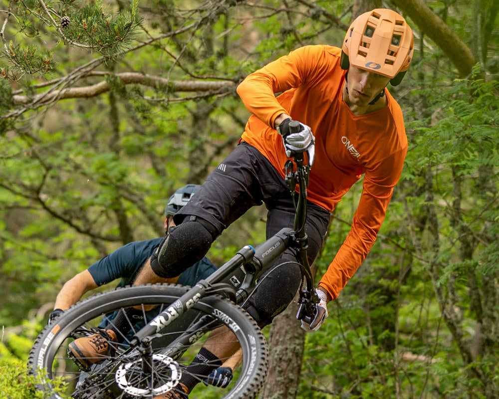 Ein Mountainbiker mit orangefarbenem Helm und Shirt fährt einen Waldweg bergab und legt sich in eine Kurve, während ihm dicht auf den Fersen ein anderer Radfahrer in dunkler Kleidung folgt.