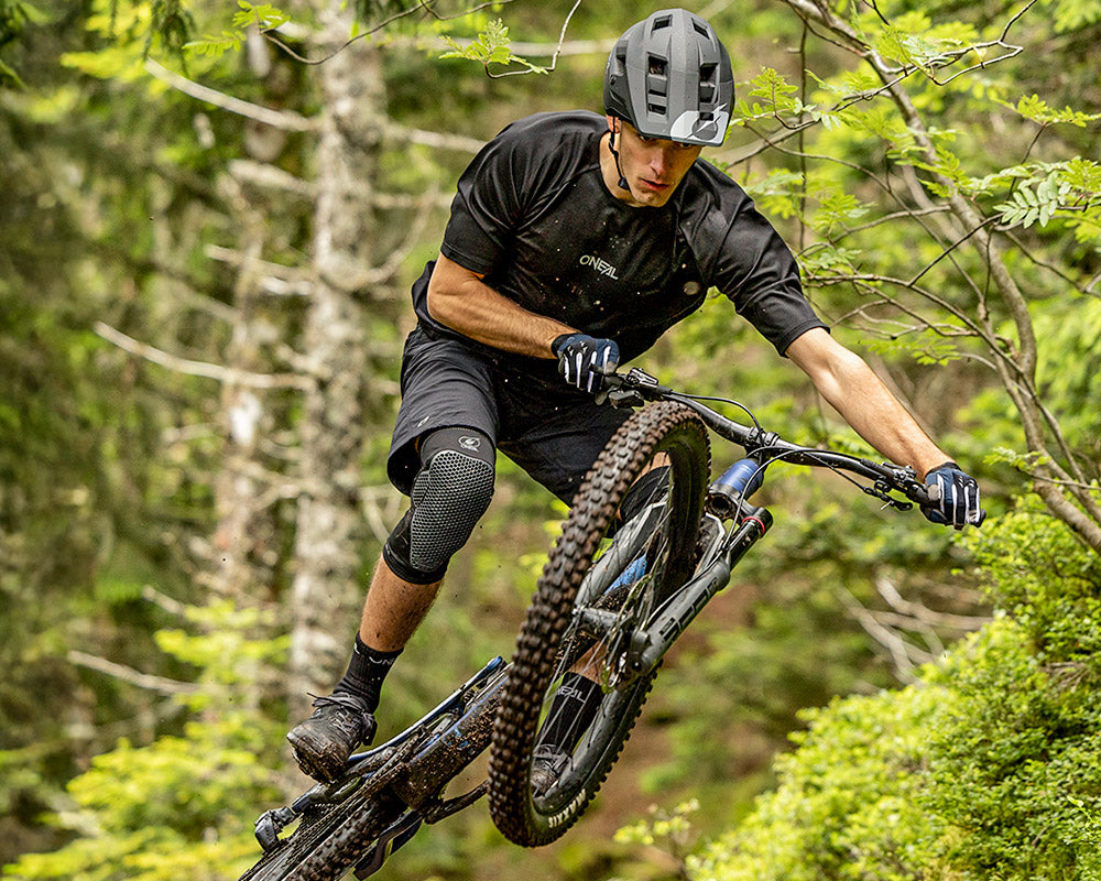 Ein Mountainbiker mit Helm und Knieschützern fährt in einem Wald von einer Sprungschanze herunter, wobei er sich mit ausgestrecktem Bein zur Seite lehnt und das Fahrrad inmitten grüner Bäume und Blätter schräg hält.