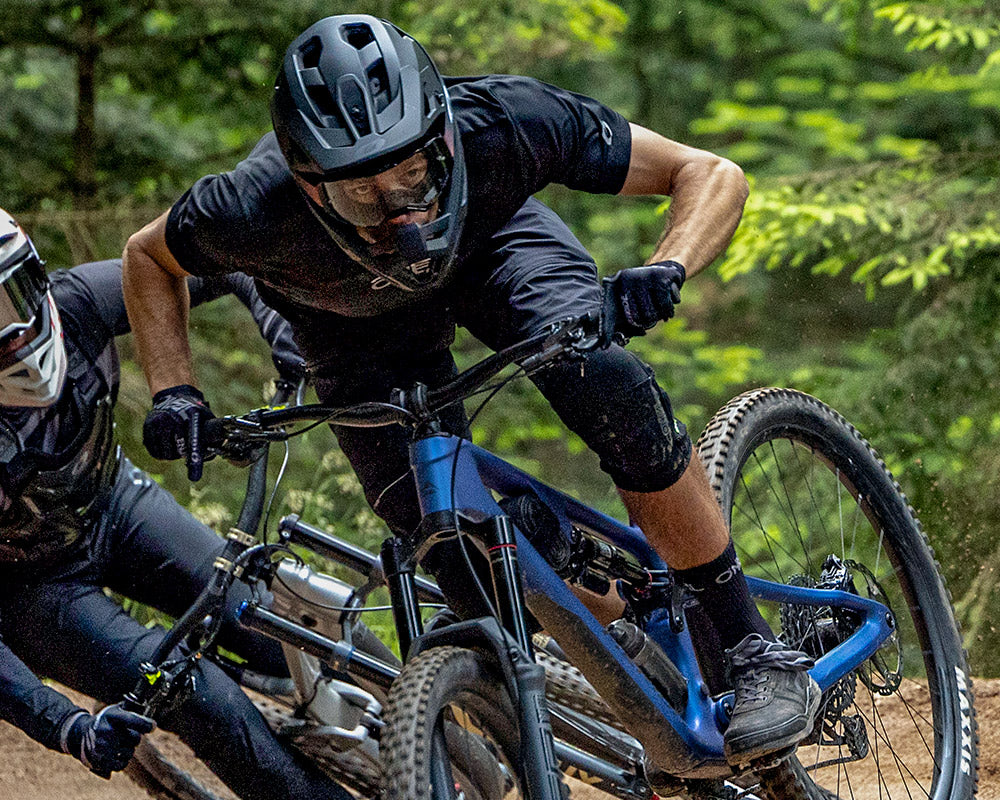 Zwei Mountainbiker mit Helmen und Schutzausrüstung rasen einen Waldweg hinunter und legen sich in eine scharfe Kurve. Bäume und Grün umgeben sie und unterstreichen die dynamische, rasante Action.