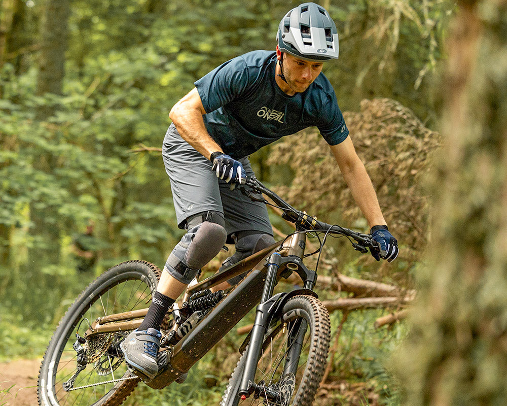 Ein Mann mit Helm und Schutzausrüstung fährt mit einem Mountainbike auf einem Feldweg durch einen Wald und legt sich mit höchster Konzentration in eine Kurve.