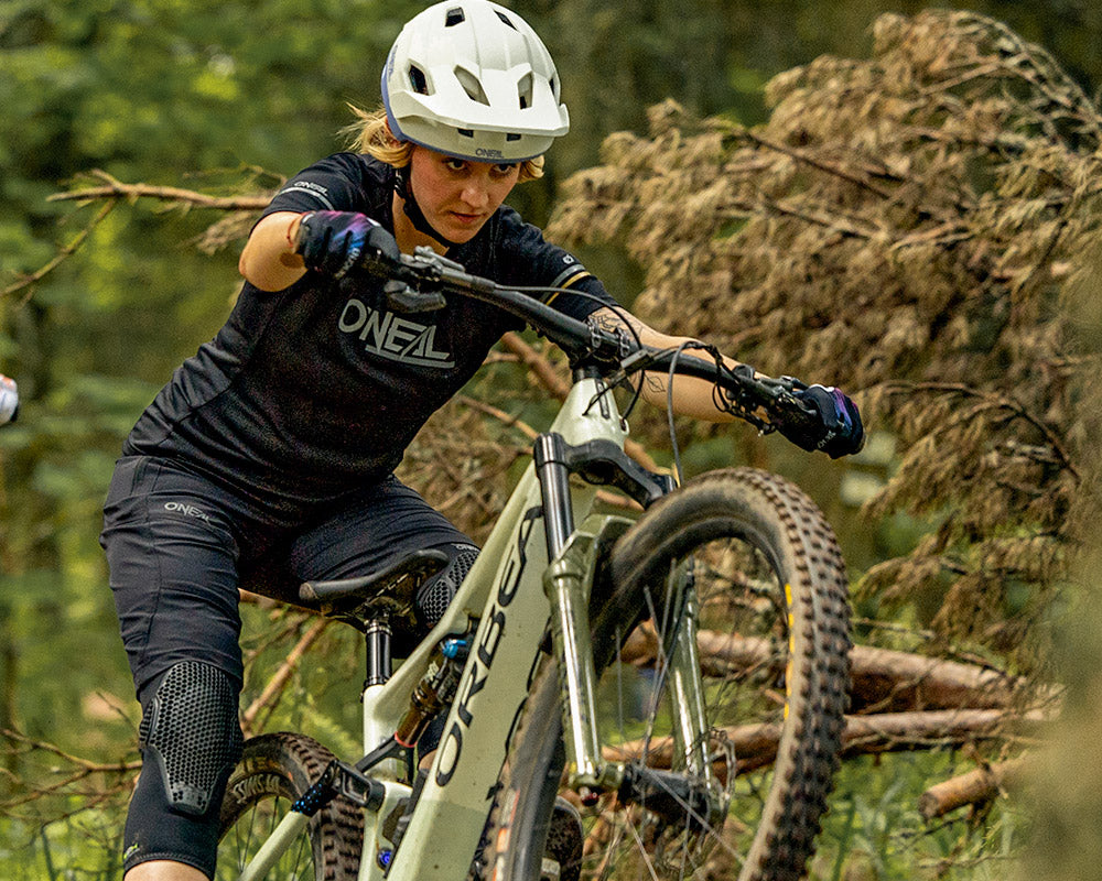 Eine Person mit Helm und Schutzausrüstung fährt mit einem Mountainbike über einen Waldweg und hebt das Vorderrad über unebenes Gelände.