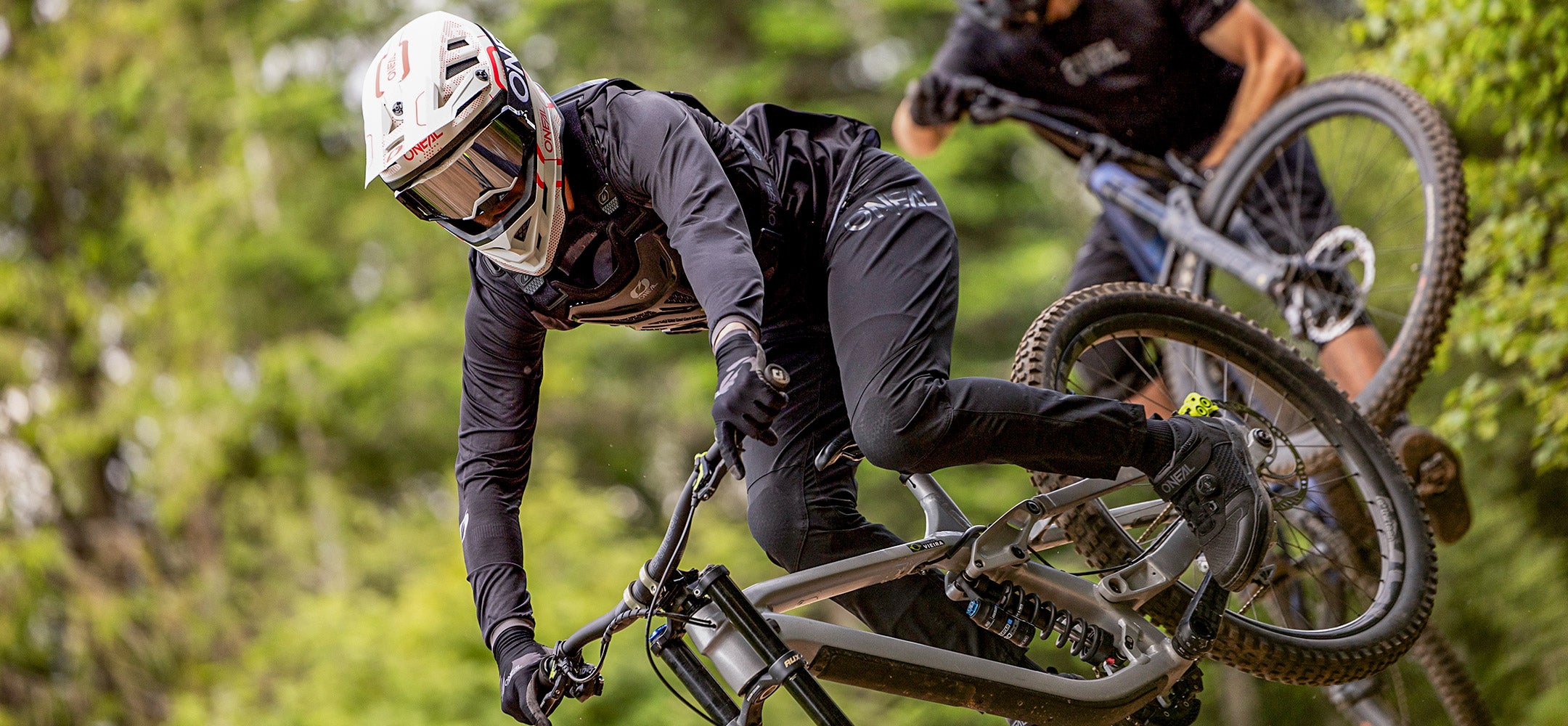 Zwei Mountainbiker in Schutzausrüstung und Helmen rasen einen Pfad durch einen Wald hinunter und legen sich scharf in eine Kurve, wobei im Hintergrund starke Fokussierung und Bewegungsunschärfe zu sehen sind.