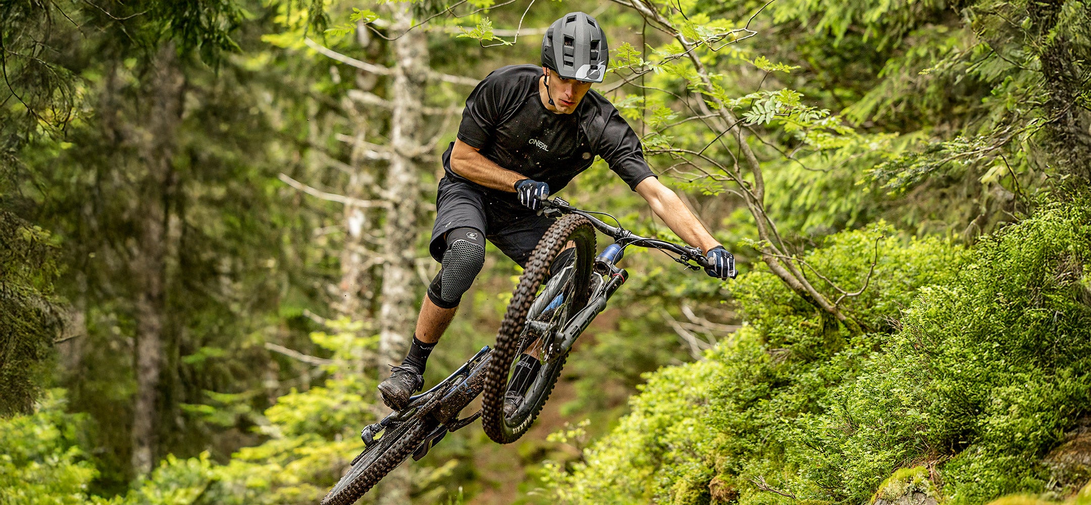 Ein Mountainbiker mit Helm und Schutzausrüstung macht einen Sprung auf einem Waldweg, umgeben von üppigen grünen Bäumen und Laub.