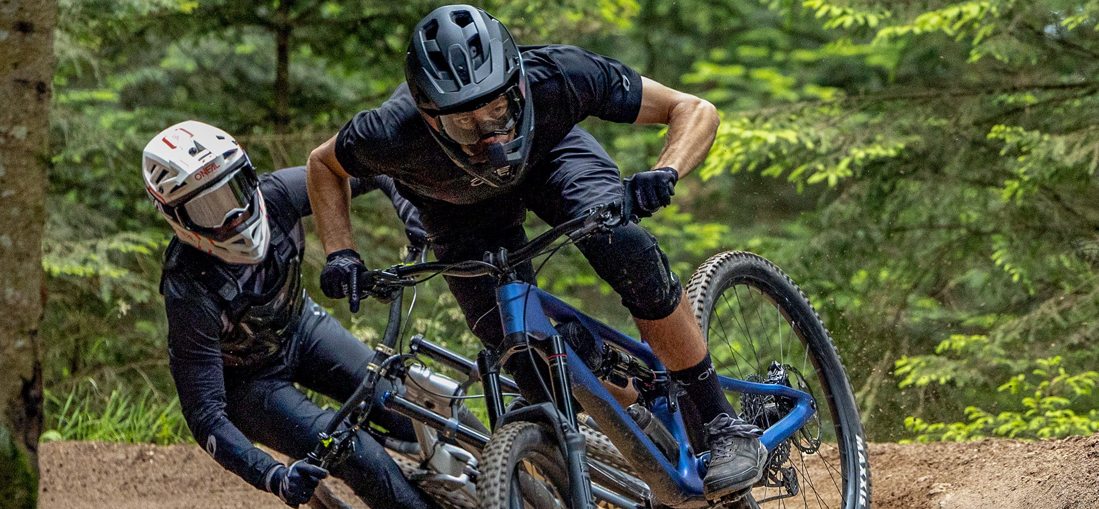 Zwei Mountainbiker mit Helmen und Schutzausrüstung rasen auf einem Feldweg durch einen Wald und legen sich scharf in eine Kurve, im Hintergrund sind Bäume und Grün zu sehen.