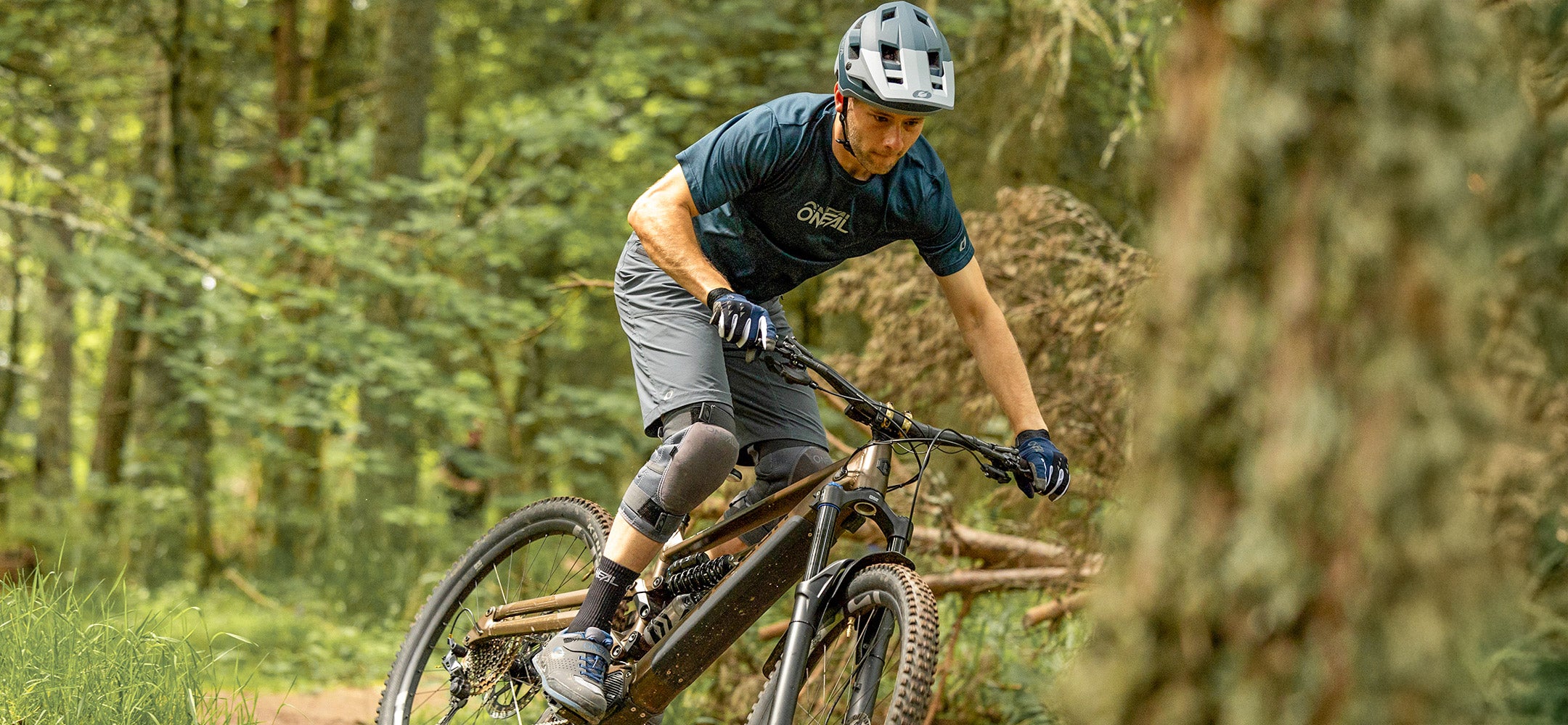 Ein Mann mit Helm und Schutzausrüstung fährt mit einem Mountainbike auf einem Waldweg und lehnt sich in eine Kurve, die von grünen Bäumen und Vegetation umgeben ist.