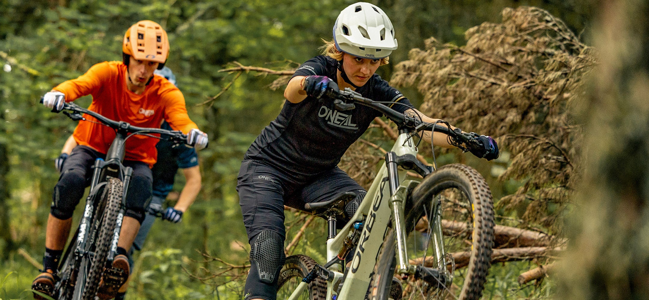 Zwei Mountainbiker mit Helmen und Schutzausrüstung fahren durch einen Waldweg, wobei der vordere Fahrer das Vorderrad seines Fahrrads über ein Hindernis hebt, während der hintere Fahrer dicht auffährt.