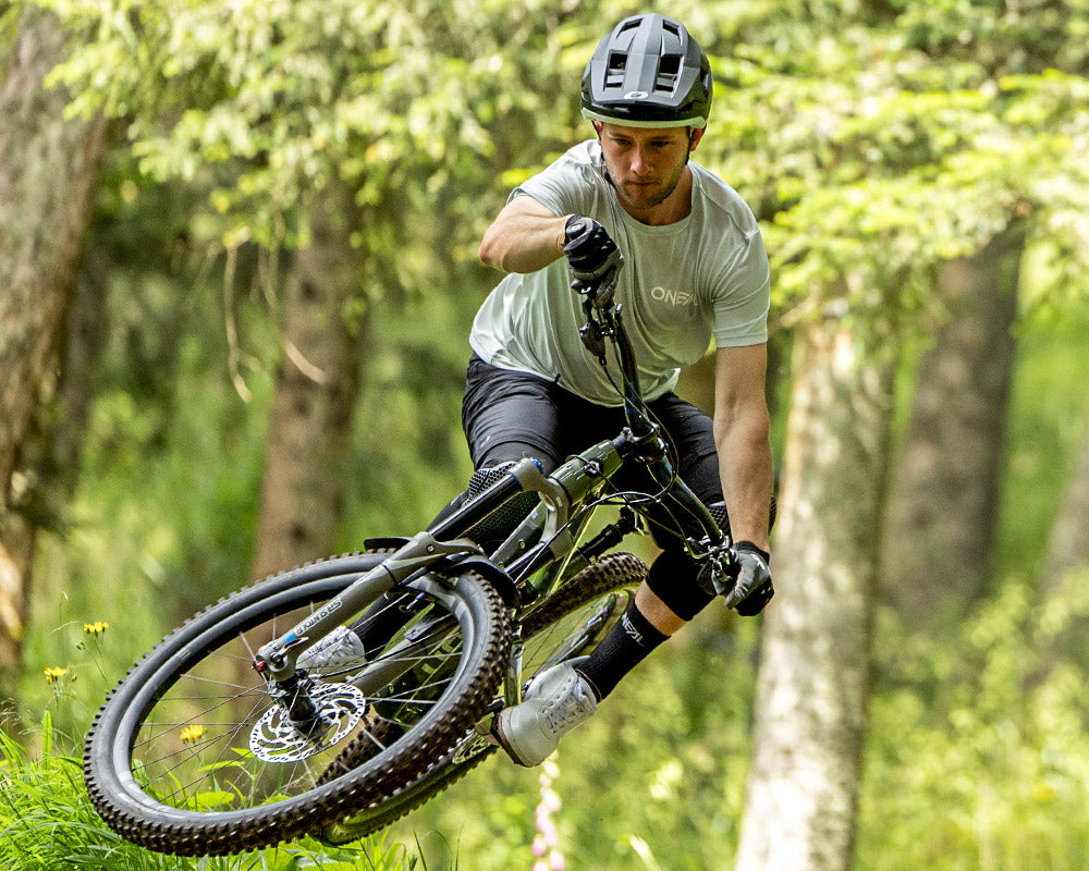 Ein Mountainbiker mit Helm und Handschuhen fährt schnell einen Waldweg hinunter und neigt sich beim Navigieren einer Kurve stark zur Seite, umgeben von grünen Bäumen und Gras.