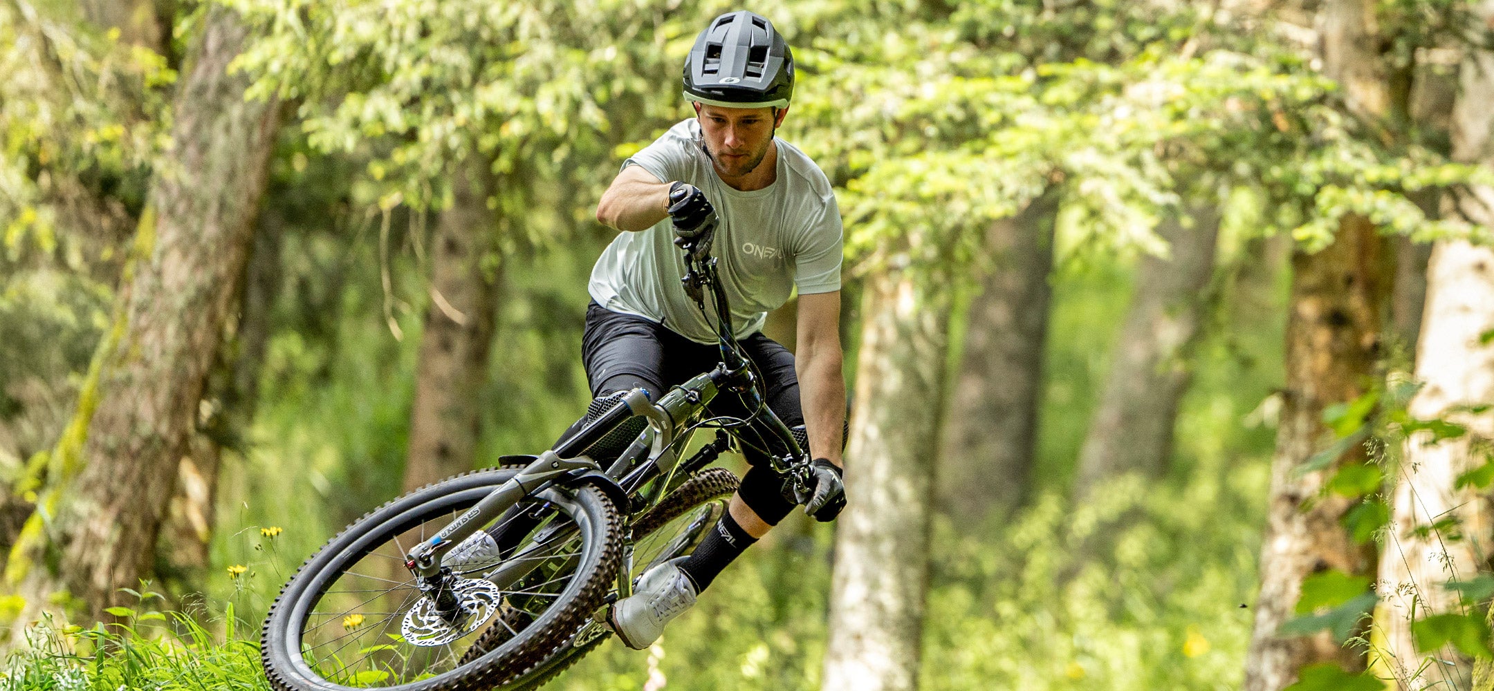 Ein Radfahrer mit Helm und Schutzausrüstung macht mit einem Mountainbike einen Sprung in einem Wald voller grüner Bäume, durch dessen Laub das Sonnenlicht fällt.